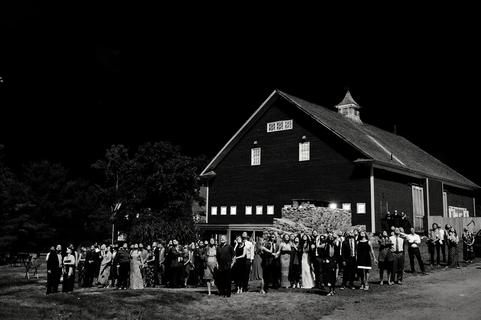 Nighttime gathering outside a large, dark barn with a group of people dressed formally, some in dresses and suits, standing on the grass in front of the barn.