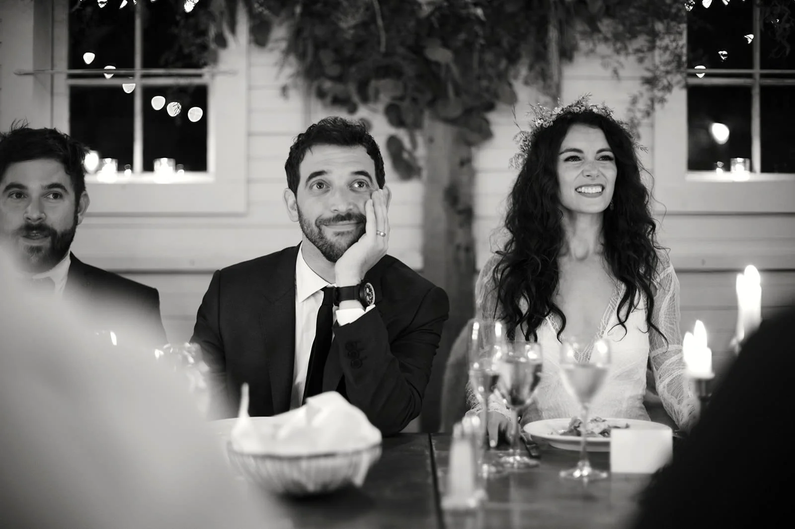 Black and white photo of a man in a suit sitting at a dinner table, resting his chin on his hand, with two women and another man in the background, candles and plates on the table, a house with windows behind them.