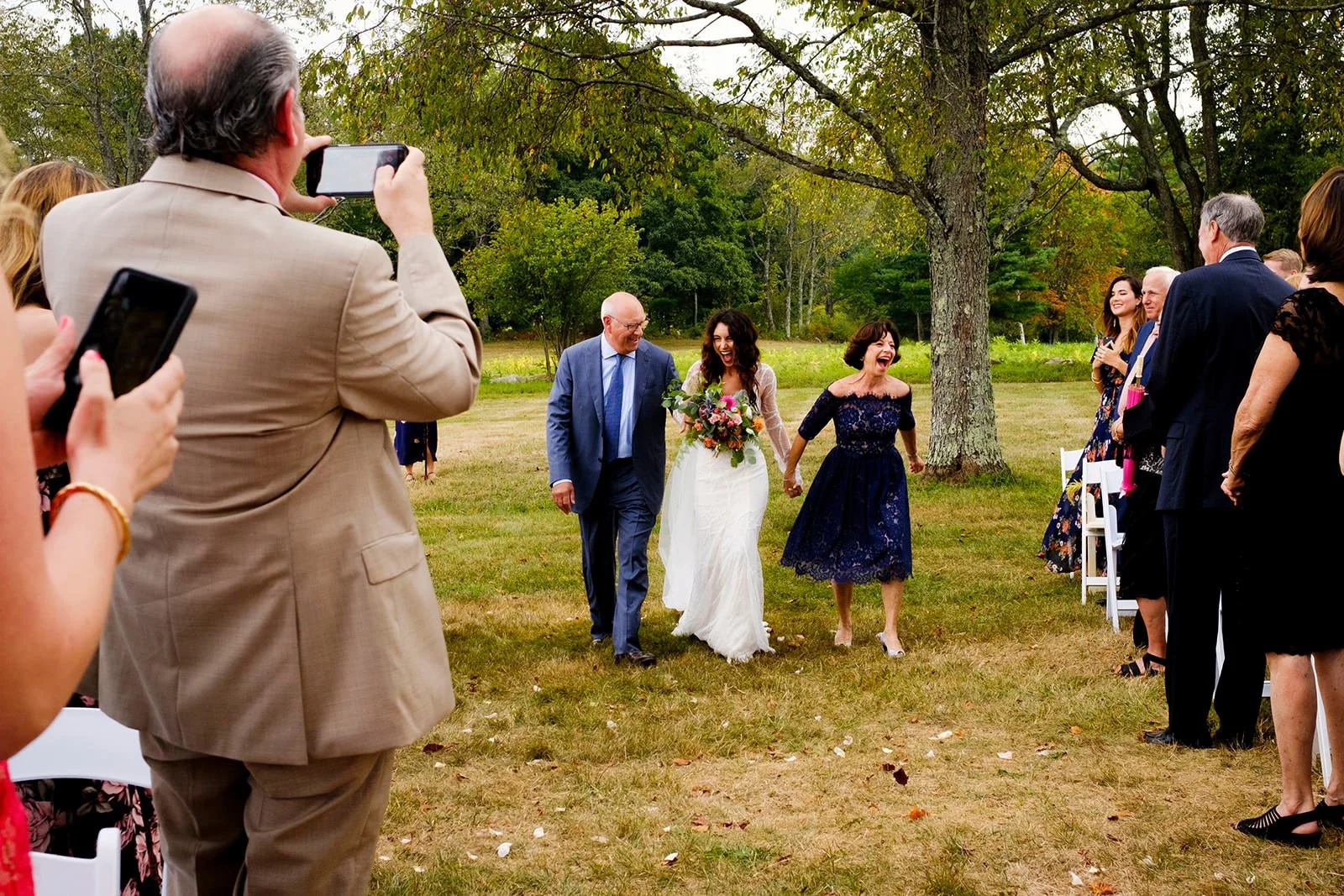 A bride holding bouquets walking down an outdoor aisle with her parents, smiling and holding hands, surrounded by guests taking photos under trees in a park-like setting.