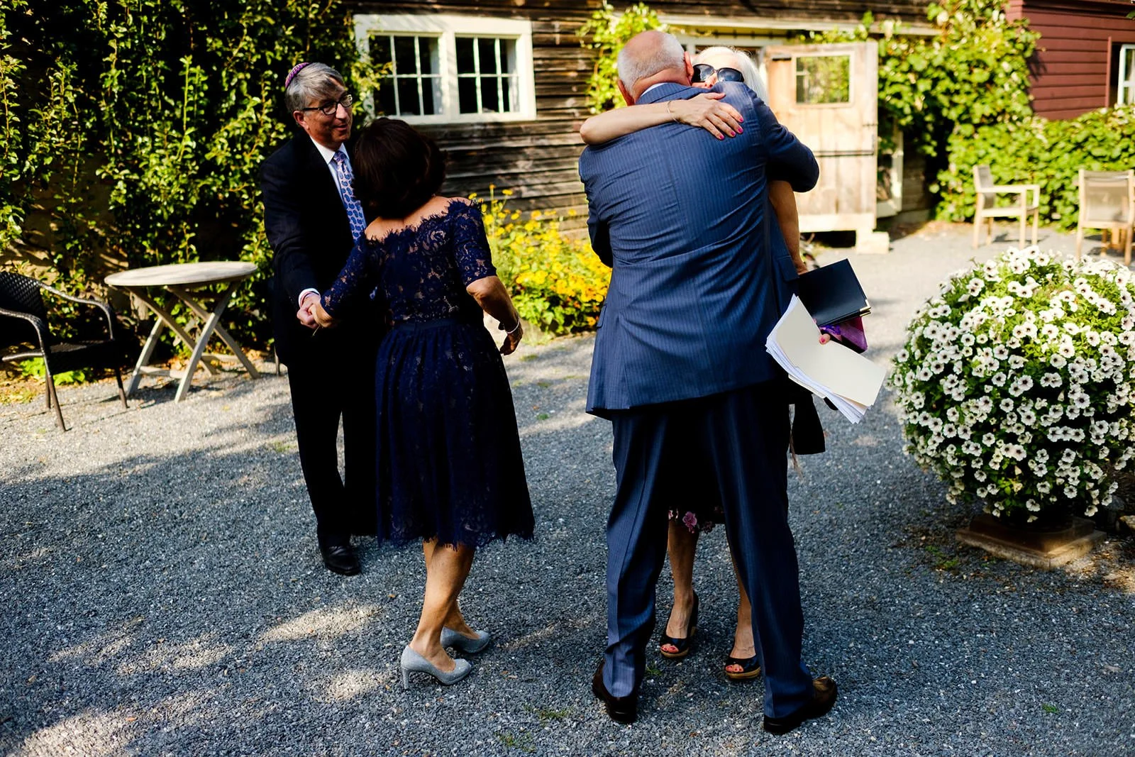A group of five people outdoors, embracing and conversing. Two women are hugging, and two men are in conversation with a woman. The setting includes a gravel ground, wooden chairs, lush greenery, and white flowers.