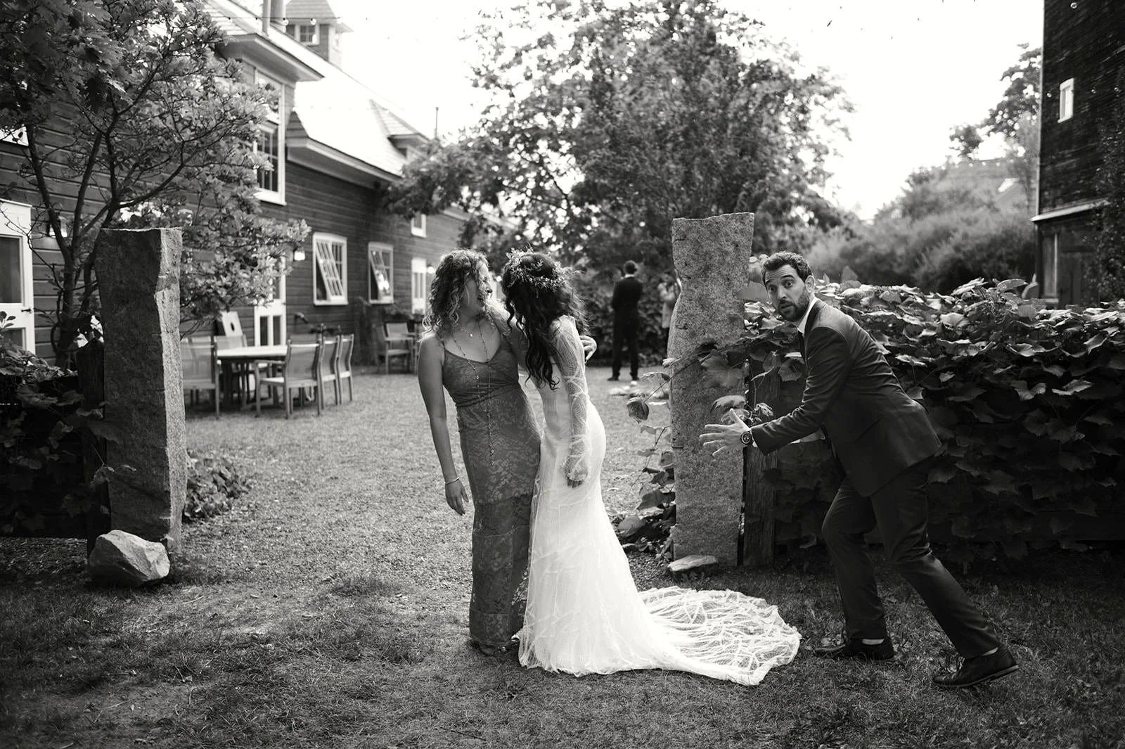 A man in a suit hiding behind a hedge and making a surprised gesture towards two women in dresses, one in a bridal gown and the other in a formal dress, at an outdoor event with buildings and trees in the background.