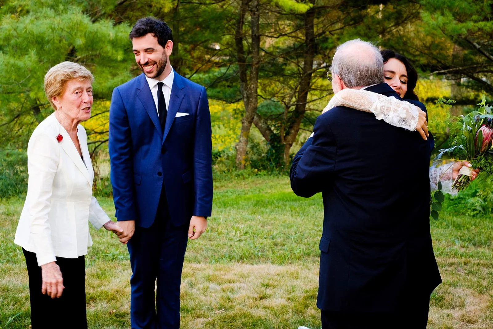 A woman in a white blazer and a woman in a dark dress hugging, while a young man in a blue suit holding hands other older man in a dark suit and glasses, outdoors in a green park.