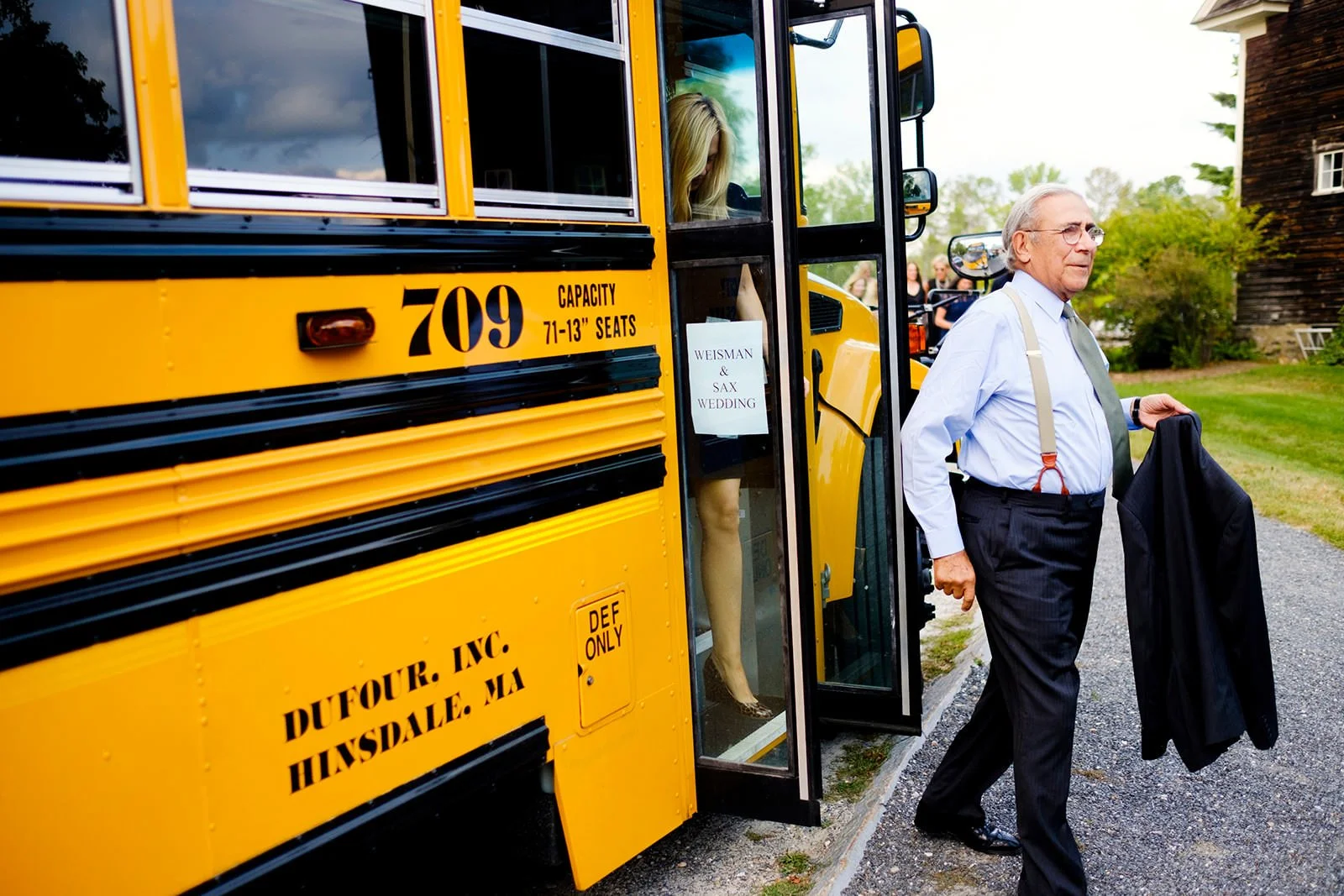 An elderly man in a white shirt, dark pants and suspenders walking away from a yellow school bus while holding a black jacket, with a woman inside the bus and a sign that reads 'WEISMAN & SAX WEDDING.' The bus is parked on a gravel path near a house 