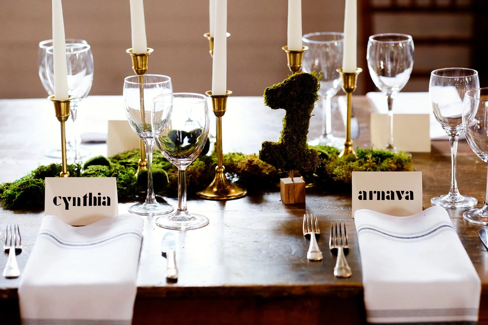A dining table set for a formal event with candles, glasses, cutlery, and table cards labeled 'Cynthia' and 'Arnav.' Green moss and a moss-covered number one centerpiece decorate the table.