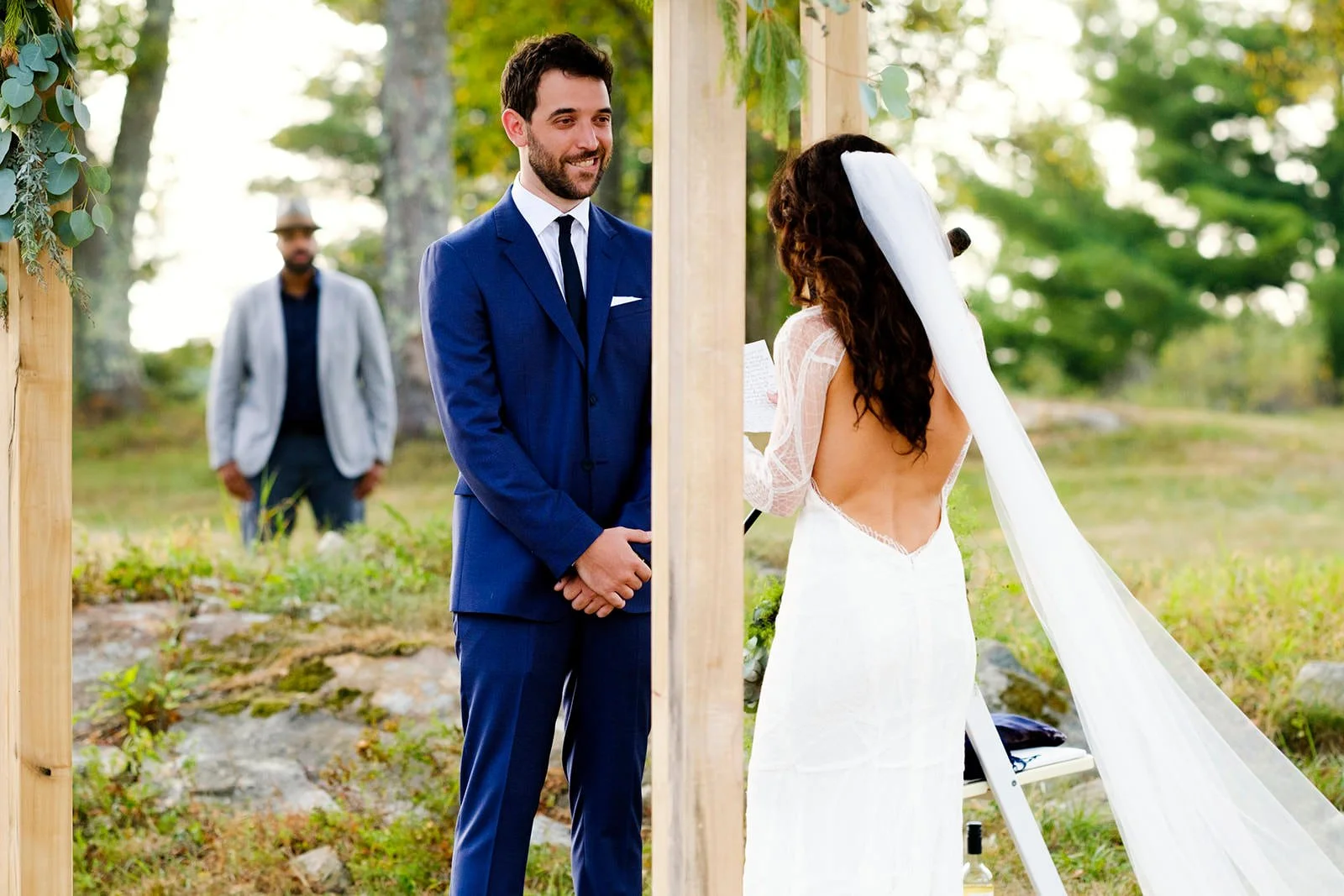 A couple gets married outdoors, with the groom smiling as he looks at the bride, who is reading her vows. The bride is wearing a white dress with an open back and a veil, while the groom is dressed in a blue suit and tie. An officiant stands nearby, 