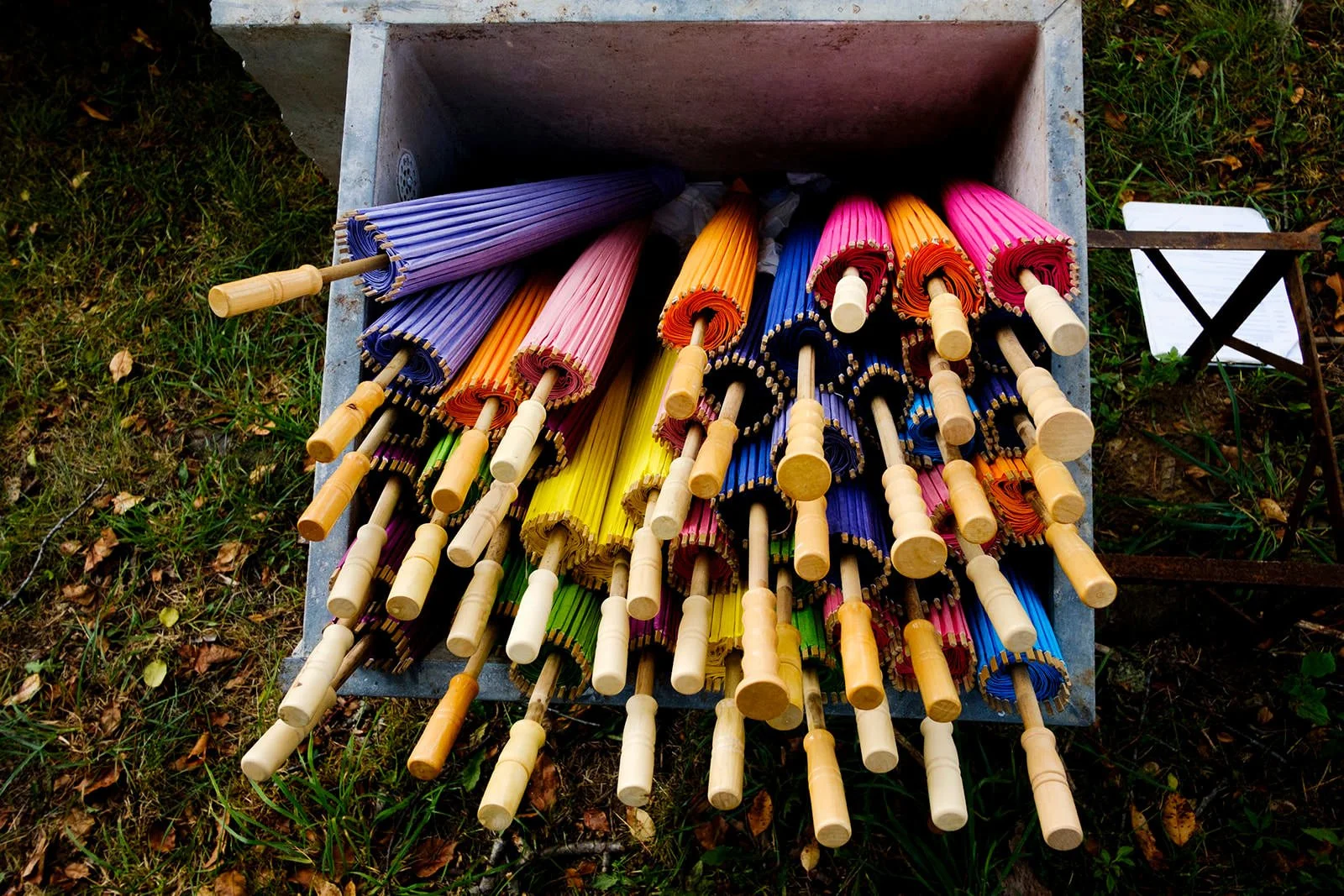 A collection of colorful umbrellas with wooden handles, stored upside down in a square container outdoors on grass.