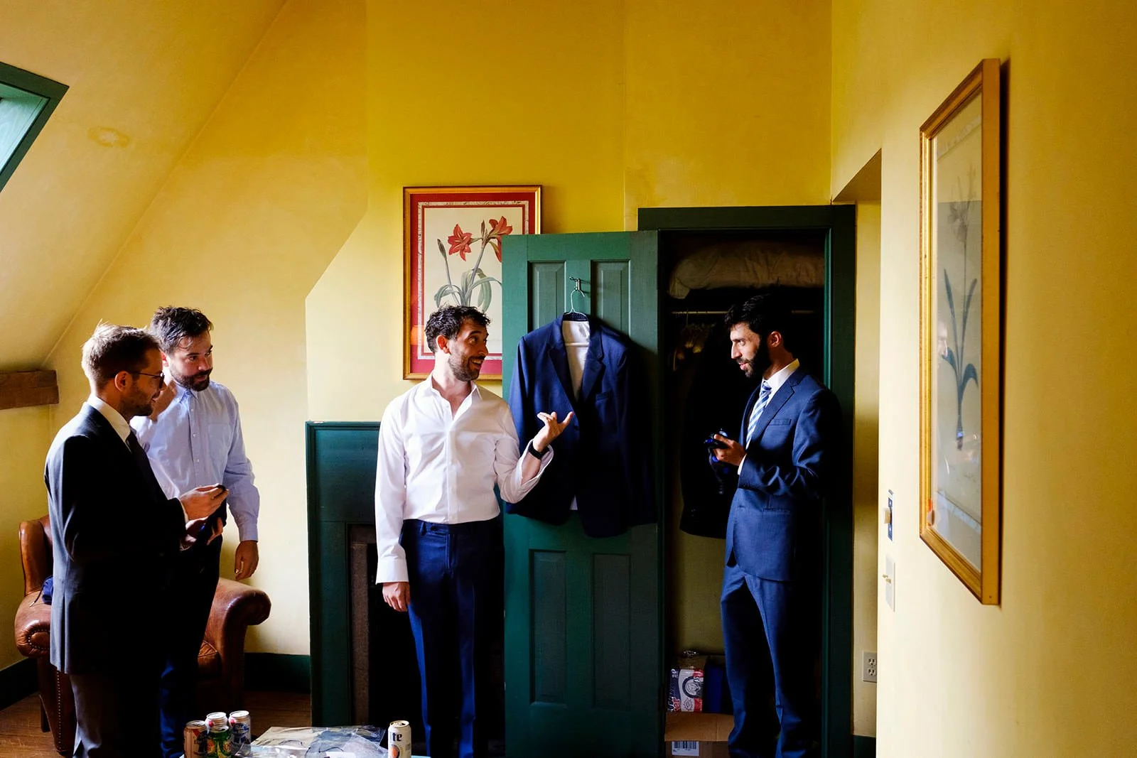 Four men in suits gathering in a room, one is showing a navy blazer to another. The room has yellow walls with framed flower artwork, a green door closet with a suit hanging inside, and a table with drinks.