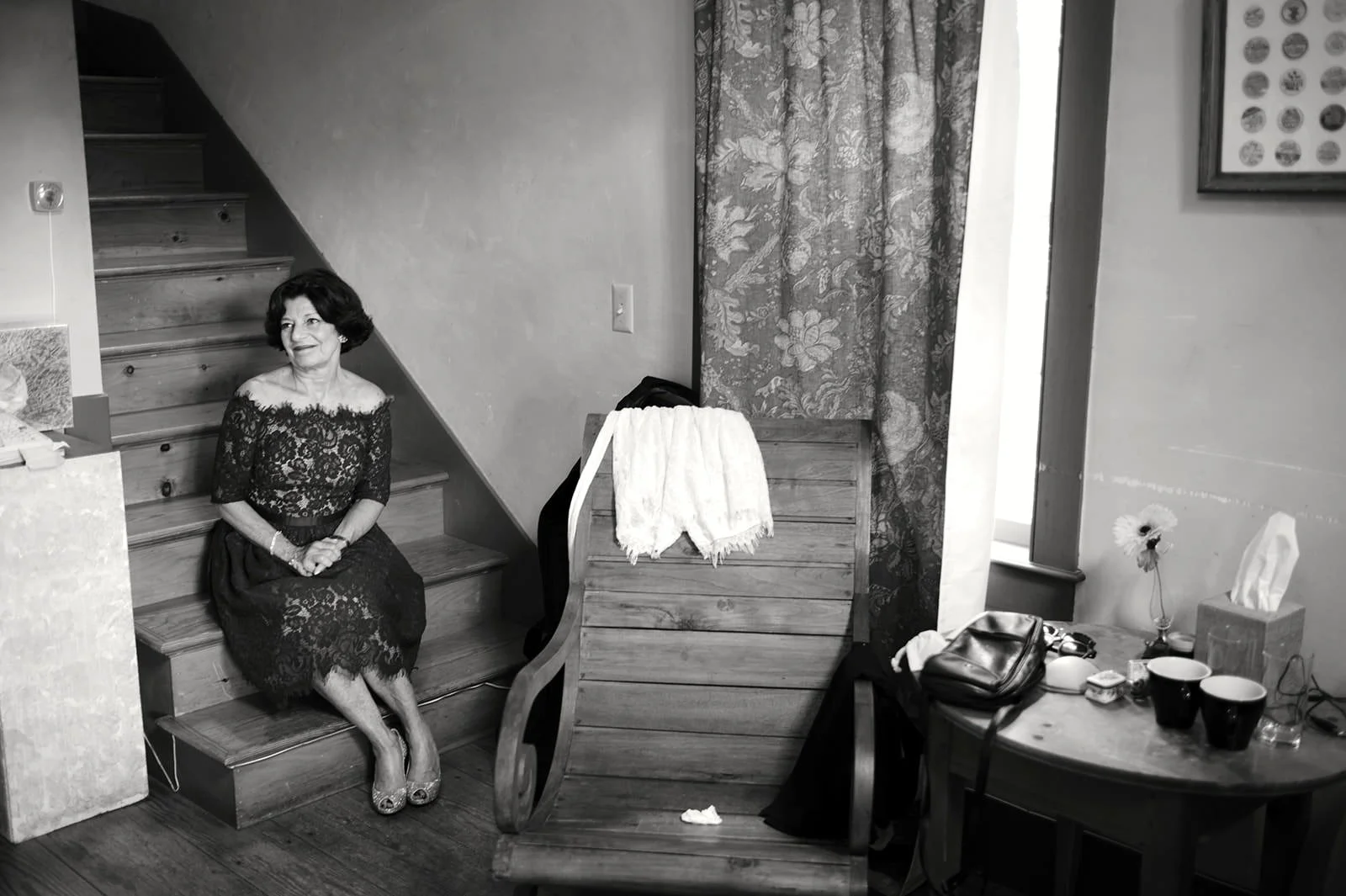 A woman sitting on stairs beside a wall, wearing a lace dress. There is a table with cups and miscellaneous items, an armchair with a towel draped over it, and a window with patterned curtains in the background.