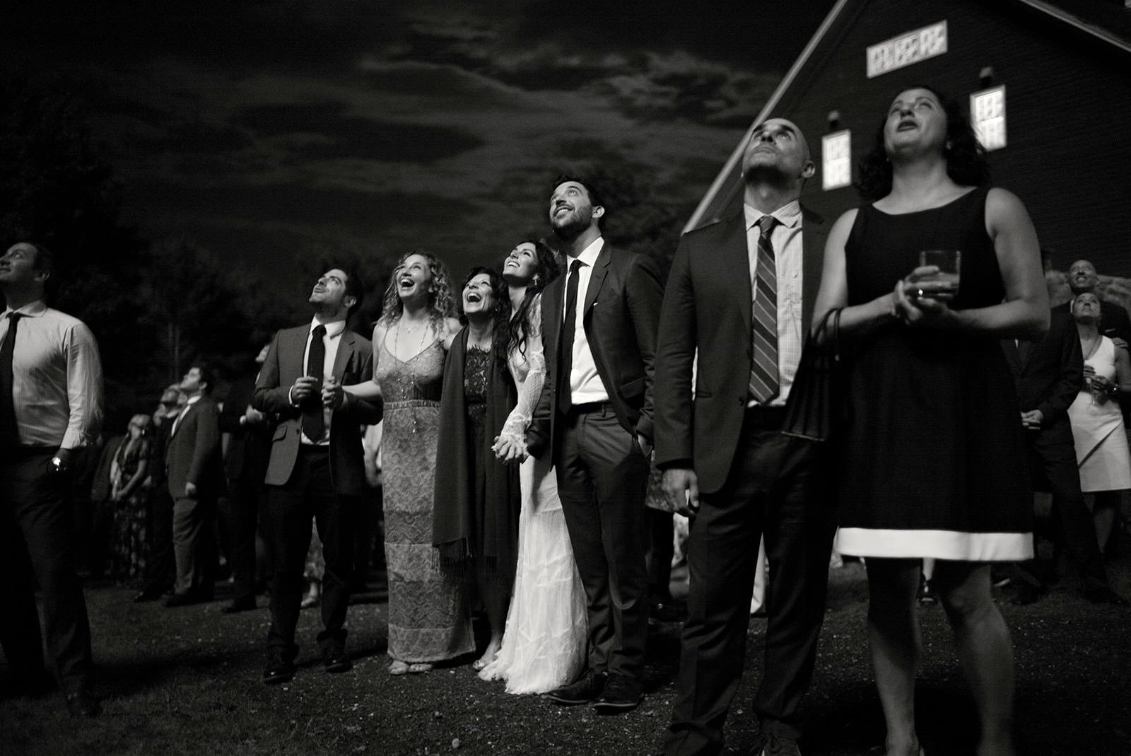 A group of people dressed in formal attire at an outdoor event at night, looking up and smiling.