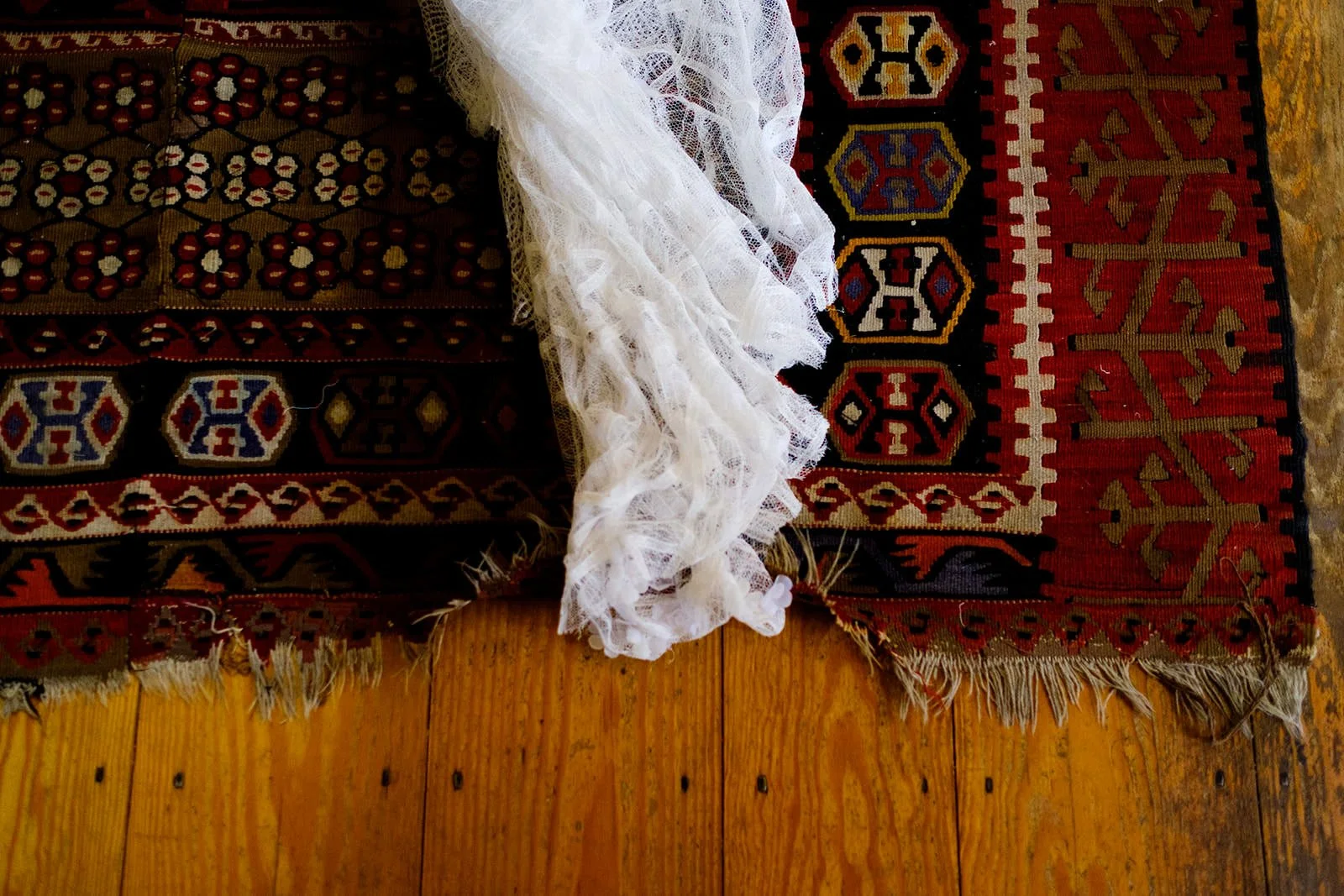 Close-up of an area of a patterned, colorful rug with a white lace fabric draped over it, all on a wooden floor.
