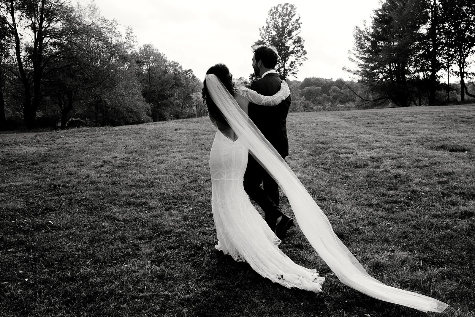 A bride and groom standing together outdoors on a grassy field, embracing, with trees in the background. The photo is in black and white.