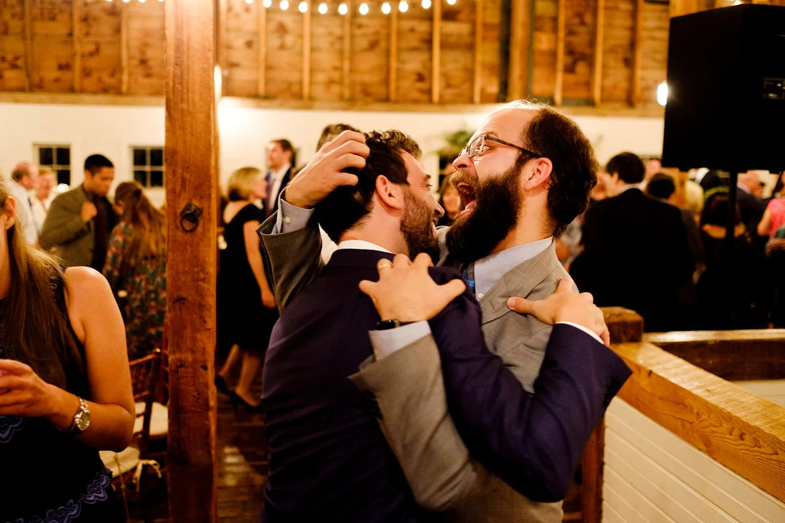 Two men dressed in suits joyfully embrace each other at a lively social event, with a crowd in the background inside a warmly-lit venue.