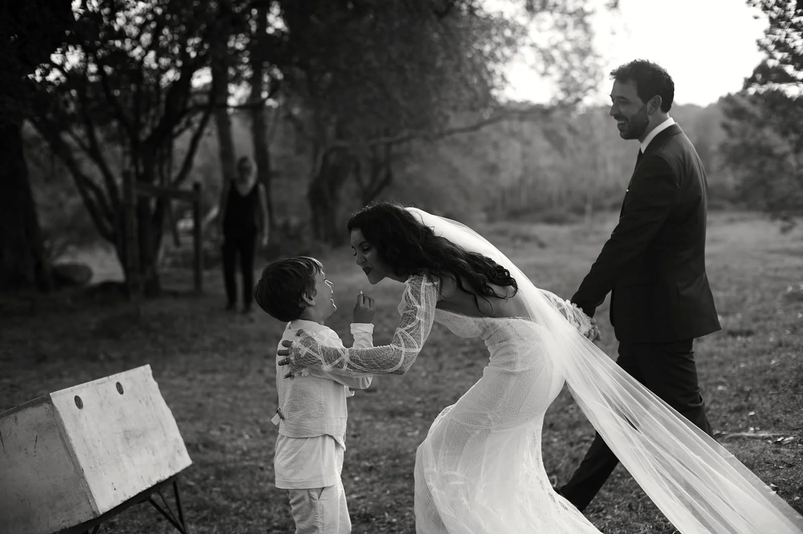A black-and-white photo of a woman in a wedding dress leaning forward to speak to a young boy, with a man in a suit holding her hand behind them, in an outdoor park setting.
