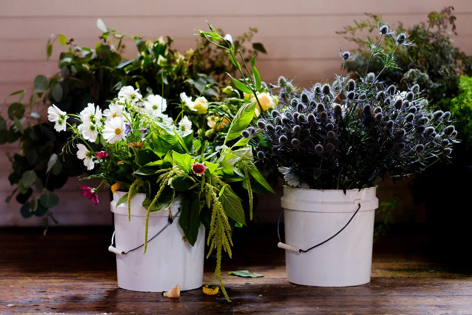 Two white buckets filled with assorted fresh flowers and green plants, sitting on a wooden surface with a wooden wall background.