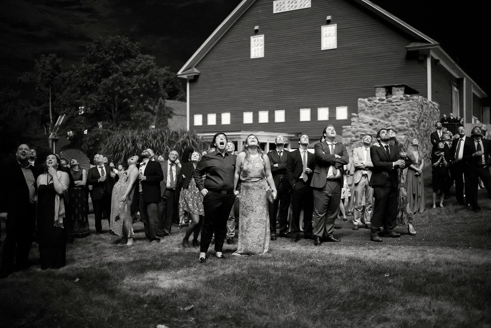 A group of people in formal attire watching an event at night outdoors, with a large house in the background.