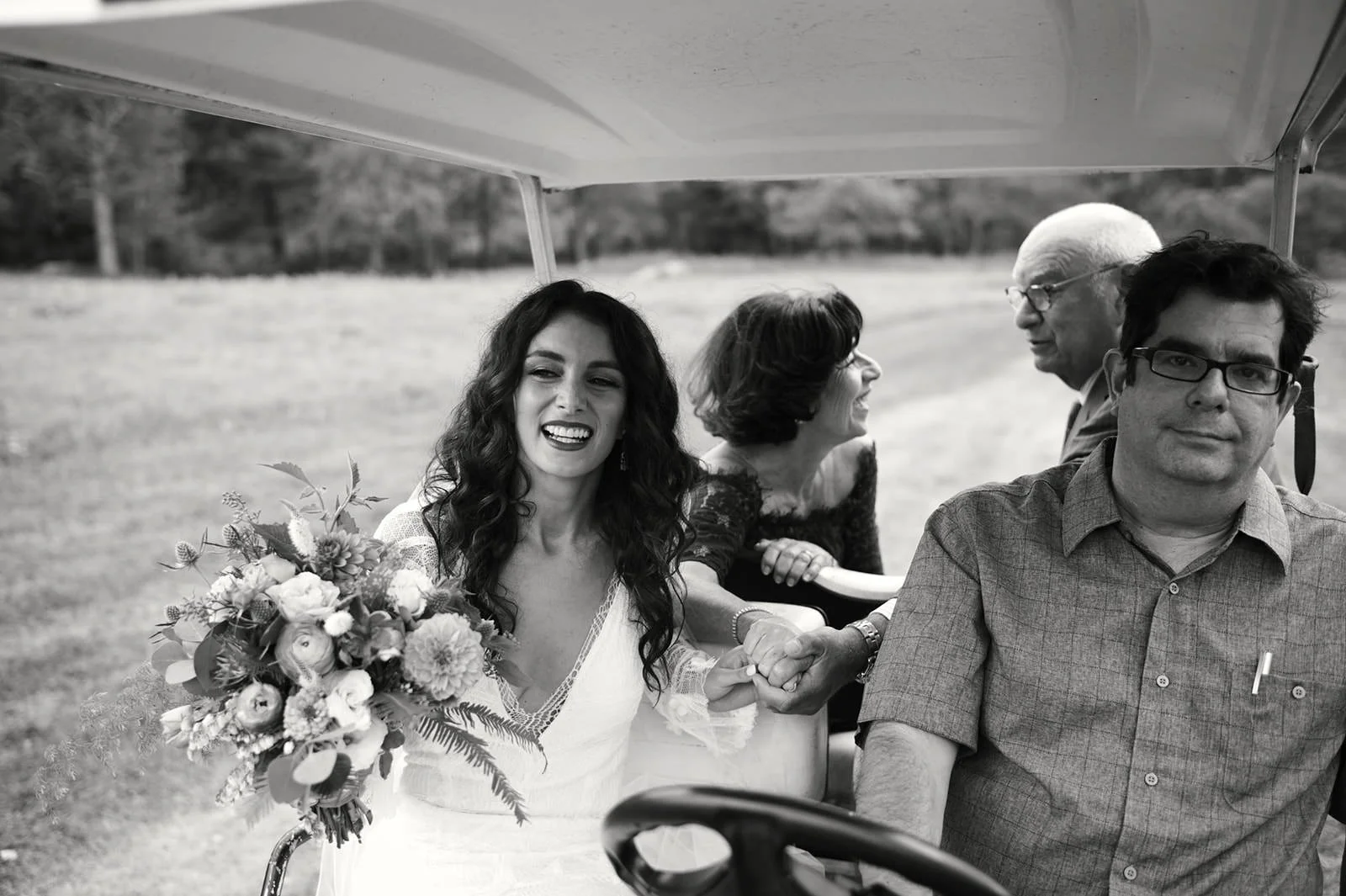 A woman holding a bouquet of flowers and smiling while holding hands with a man in a golf cart, with two women sitting behind her, outdoors on a grassy area.
