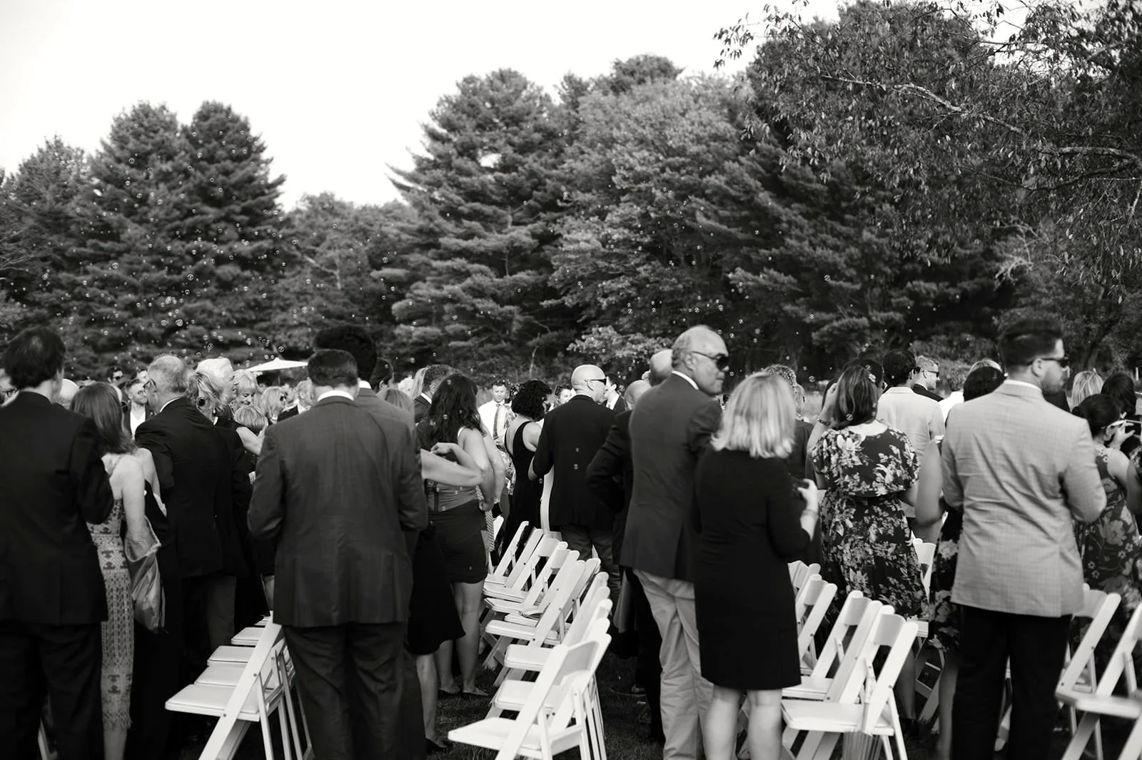 People attending an outdoor event, standing next to white chairs, with trees in the background, captured in black and white.