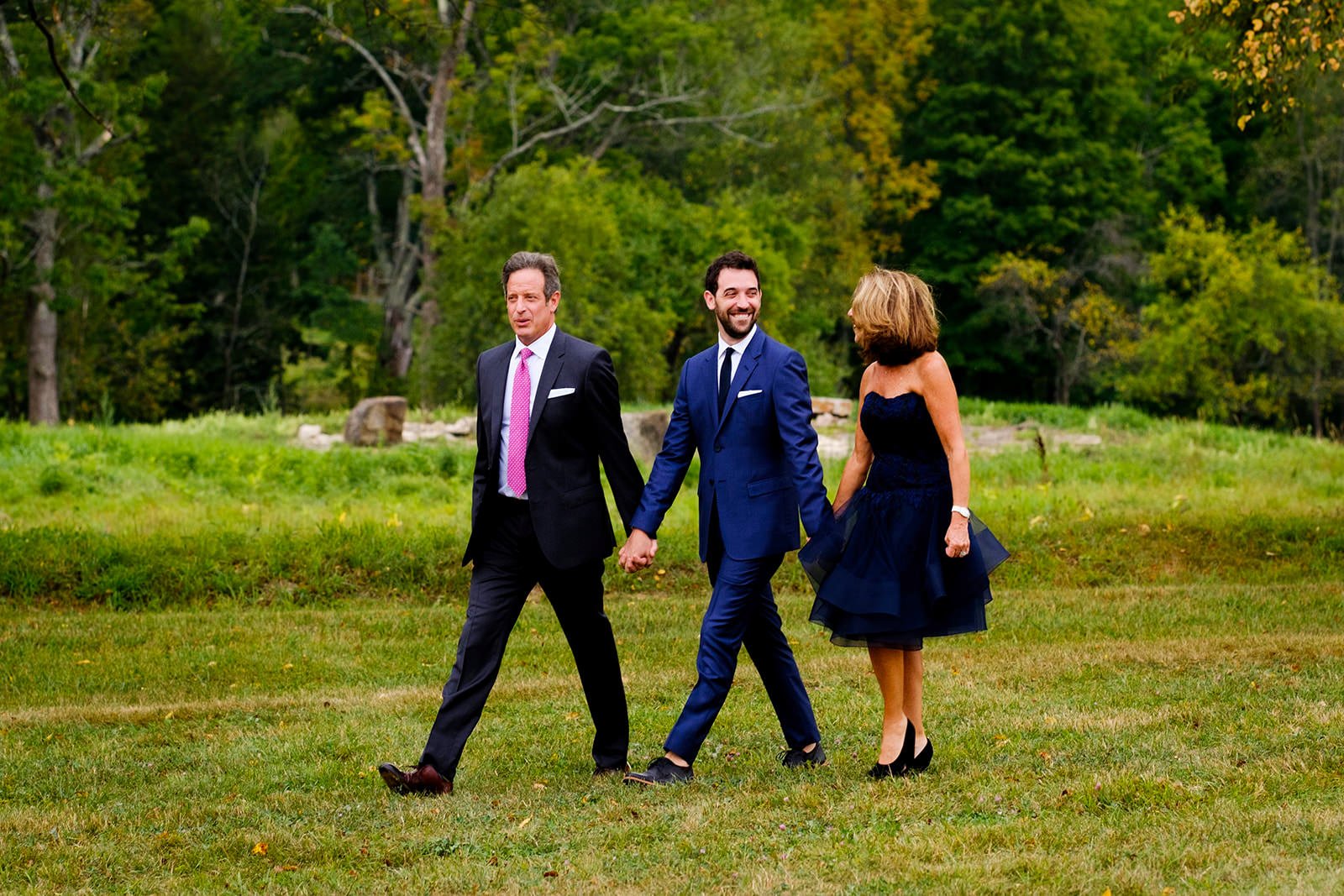 Three adults, two men and one woman, walking hand in hand outdoors on a grassy field with trees in the background. They are dressed in formal attire, with the men in suits and the woman in a strapless, knee-length navy dress. The person on the left i