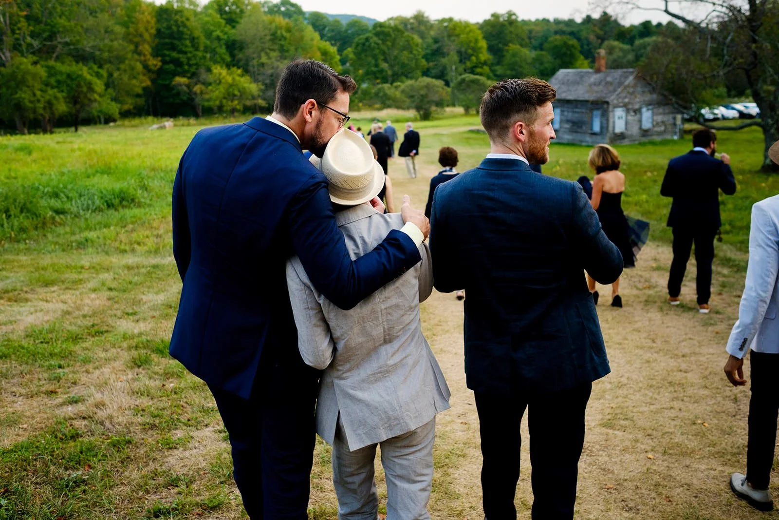 Group of people dressed in formal attire walking outdoors on a grassy path, with some people in the background near a rustic building and surrounded by trees.