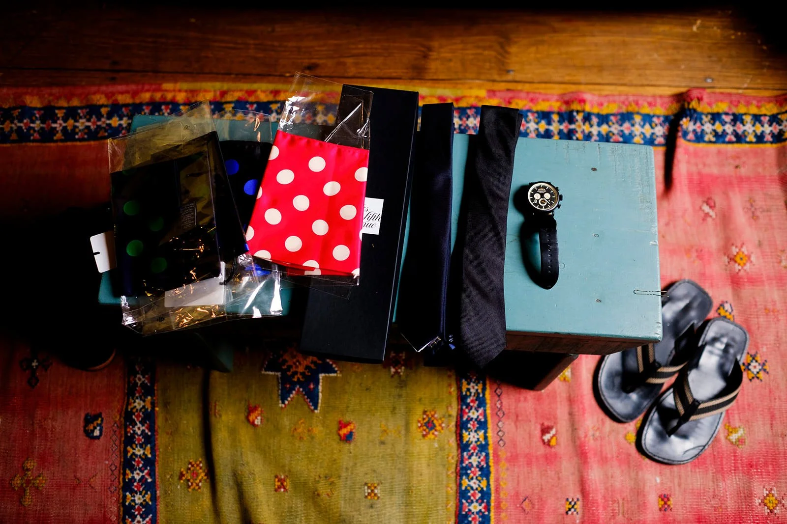 Top-down view of a light blue wooden table with a black watch, two black straps, two black surfaces, and a stack of three gift bags with polka dot designs, on a colorful patterned rug.