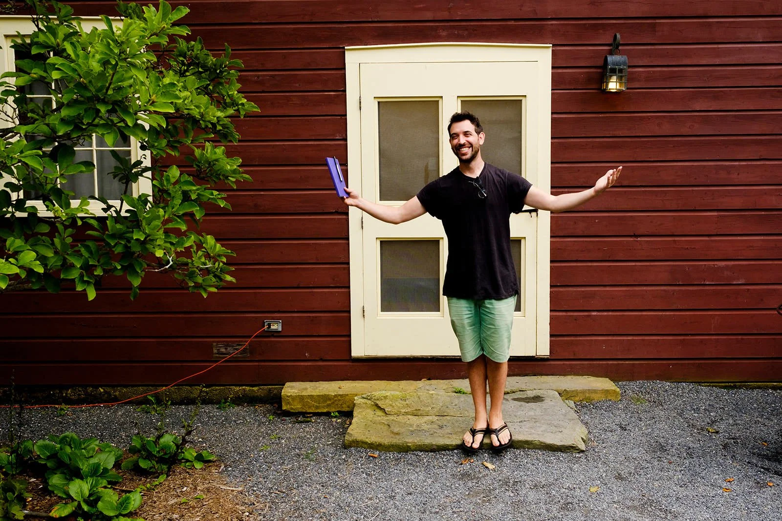 A smiling man standing on a stone step in front of a red wooden house door with glass panes, holding a tablet in one hand and arms outstretched.