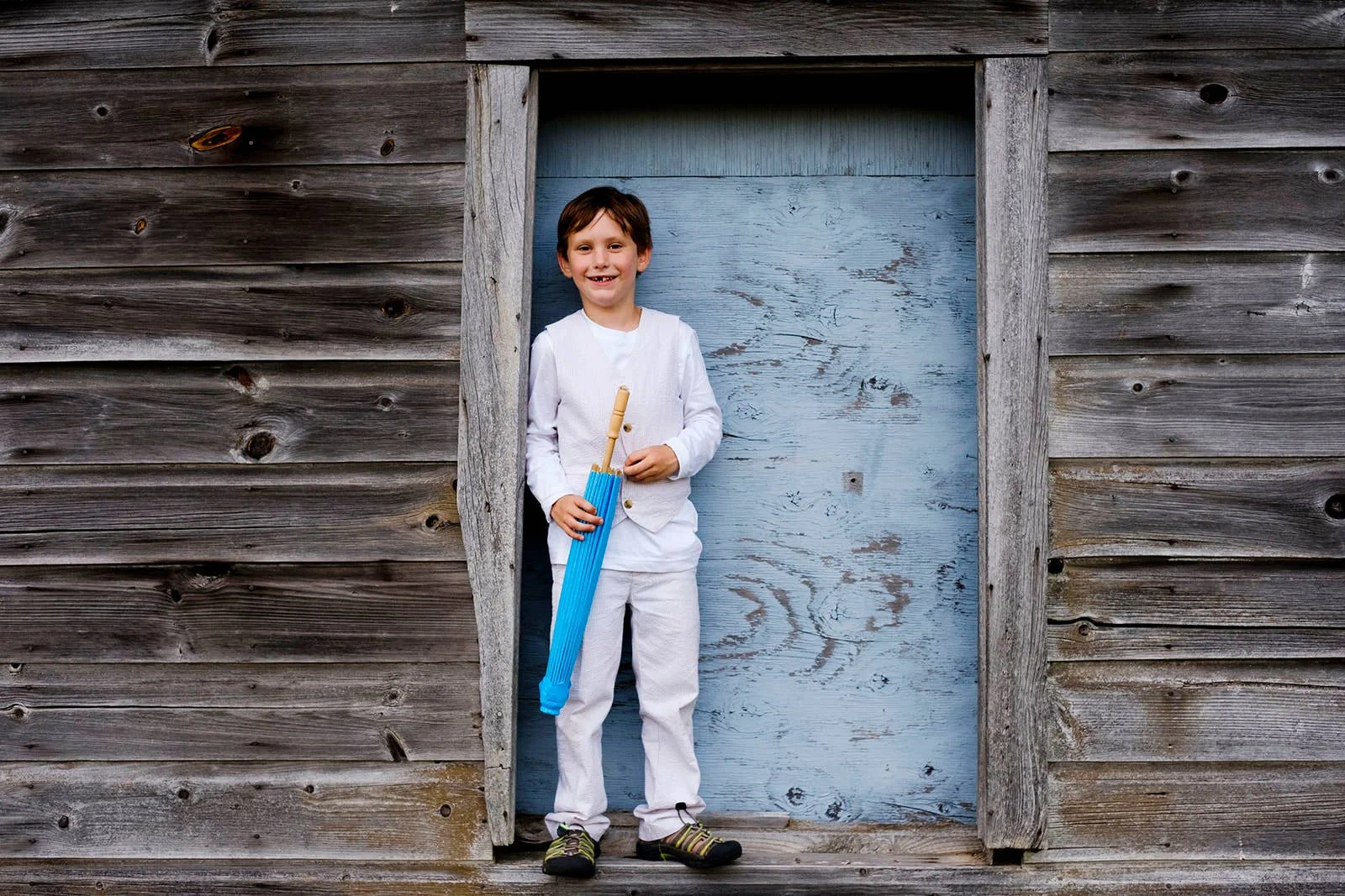 A young boy standing in a wooden doorway, holding a toy blue umbrella, smiling, wearing a white outfit and black sandals with green stripes.