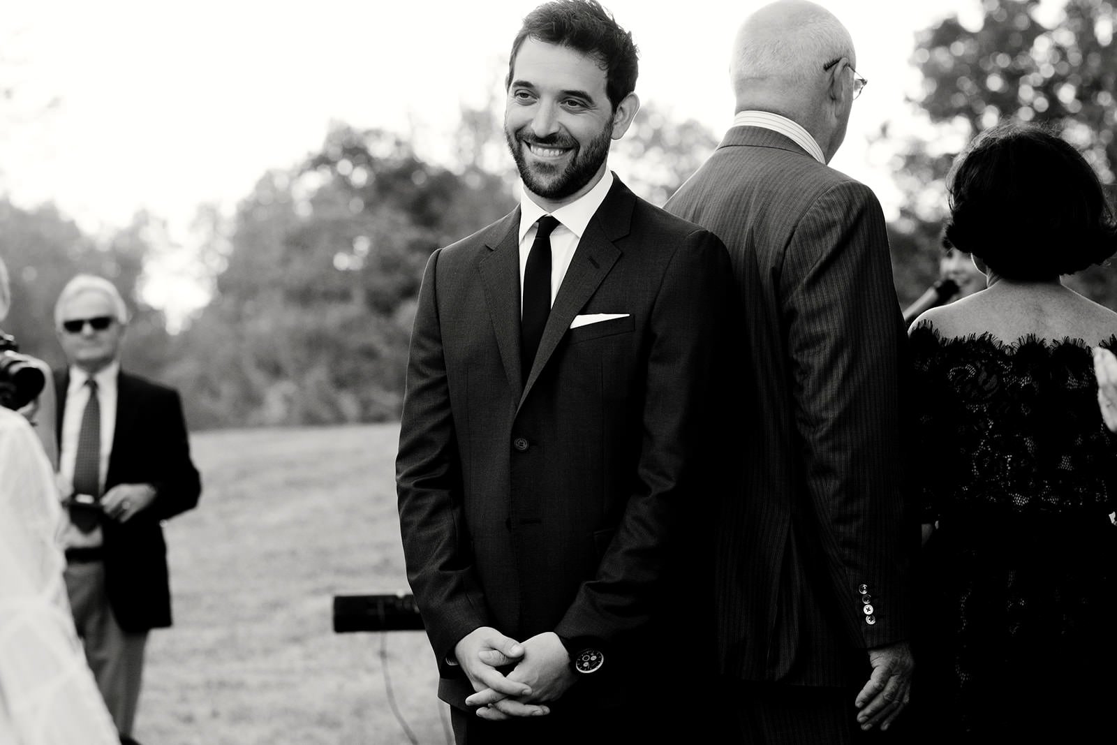 A man in a black suit with a white shirt and black tie smiling at an outdoor wedding ceremony. Others, including a woman in a black dress and men in suits, are also present.