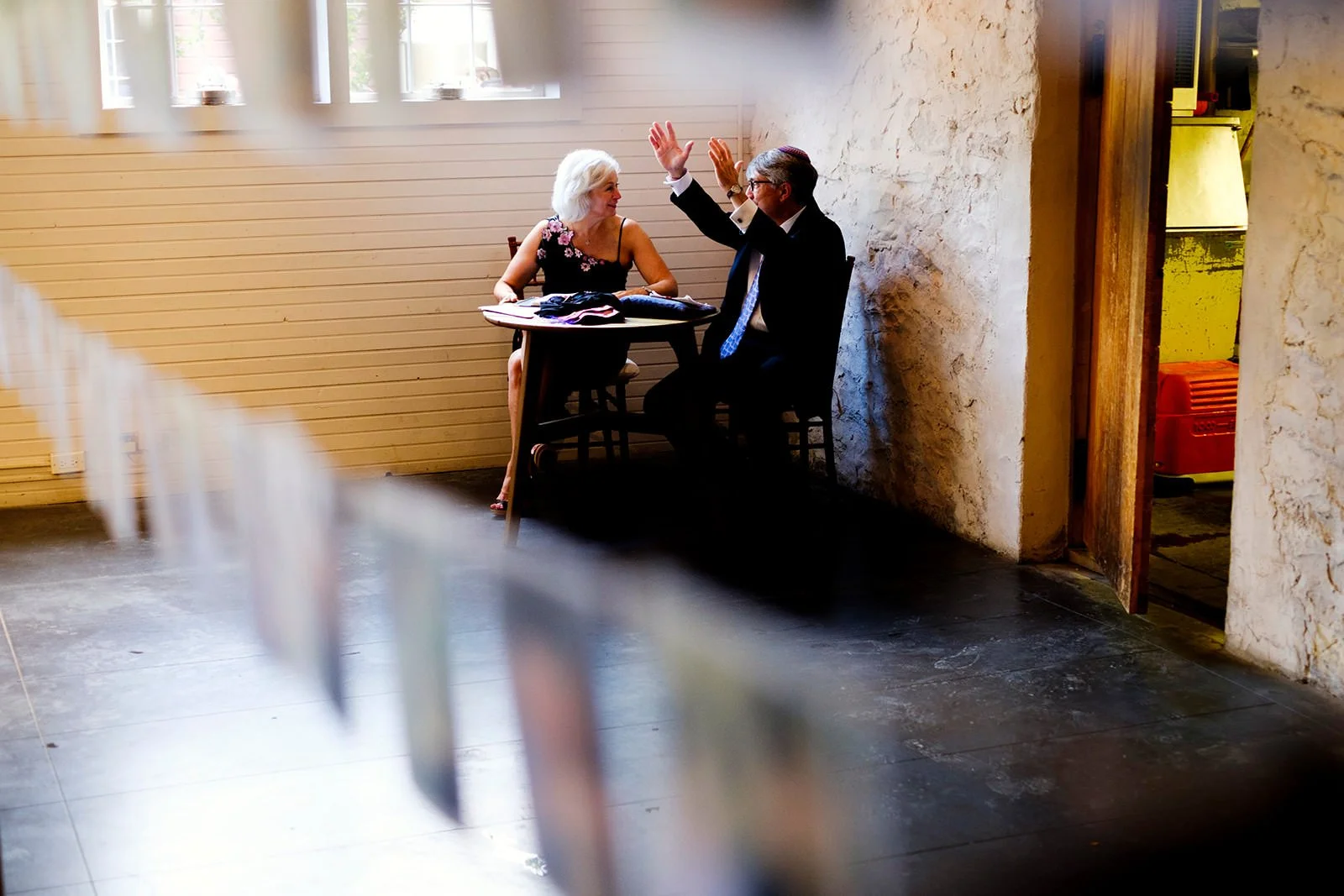 An elderly woman and an elderly man sitting at a small table in a rustic room, having a conversation and gesturing with their hands, with sunlight coming through the windows.