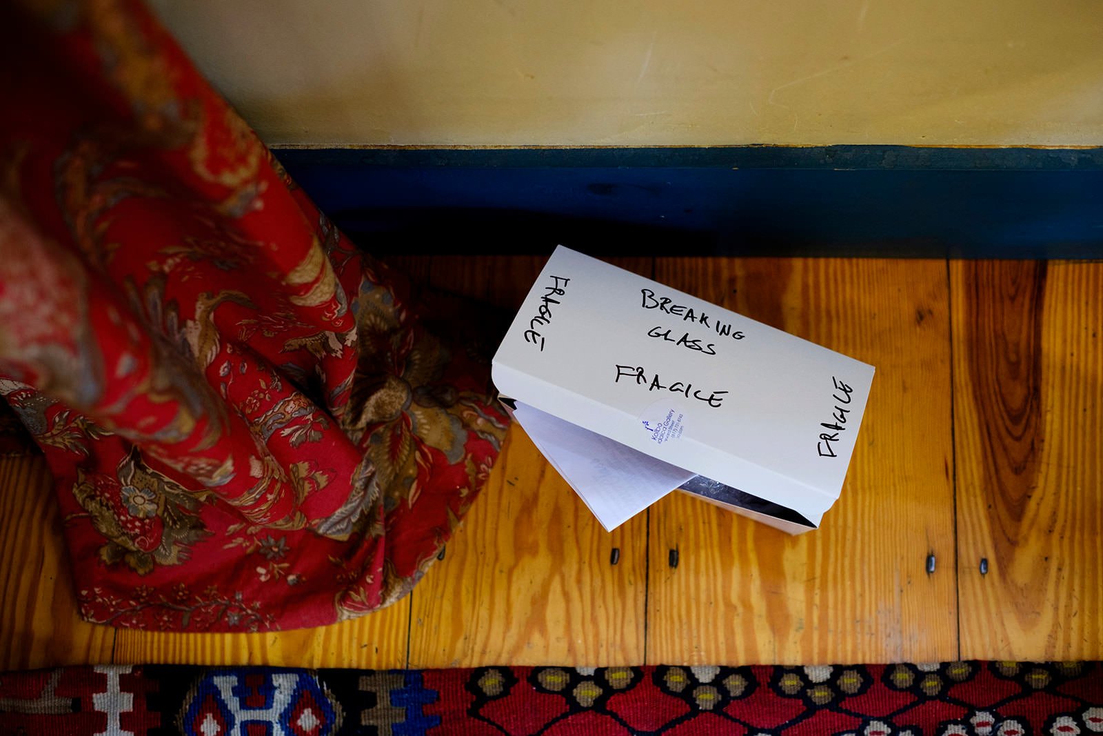 A white cardboard box labeled 'Breaking Glass Fragile' placed on a wooden surface next to a colorful fabric bag.