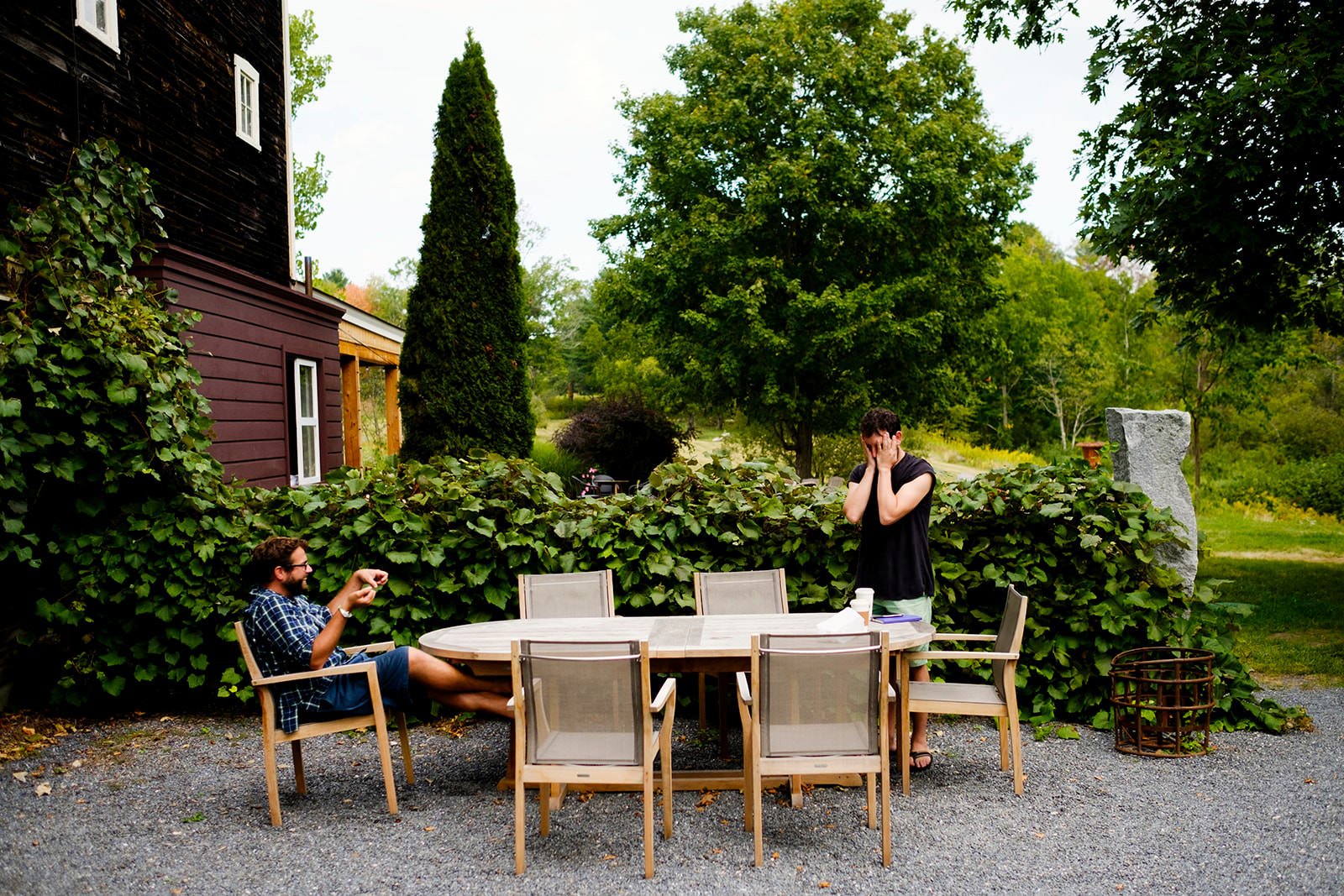 A man sitting at a round outdoor table talking on the phone, while another man wearing sunglasses and a plaid shirt is seated nearby, gesturing with his hand. The setting is a garden with lush green trees and bushes, and a red building with white win
