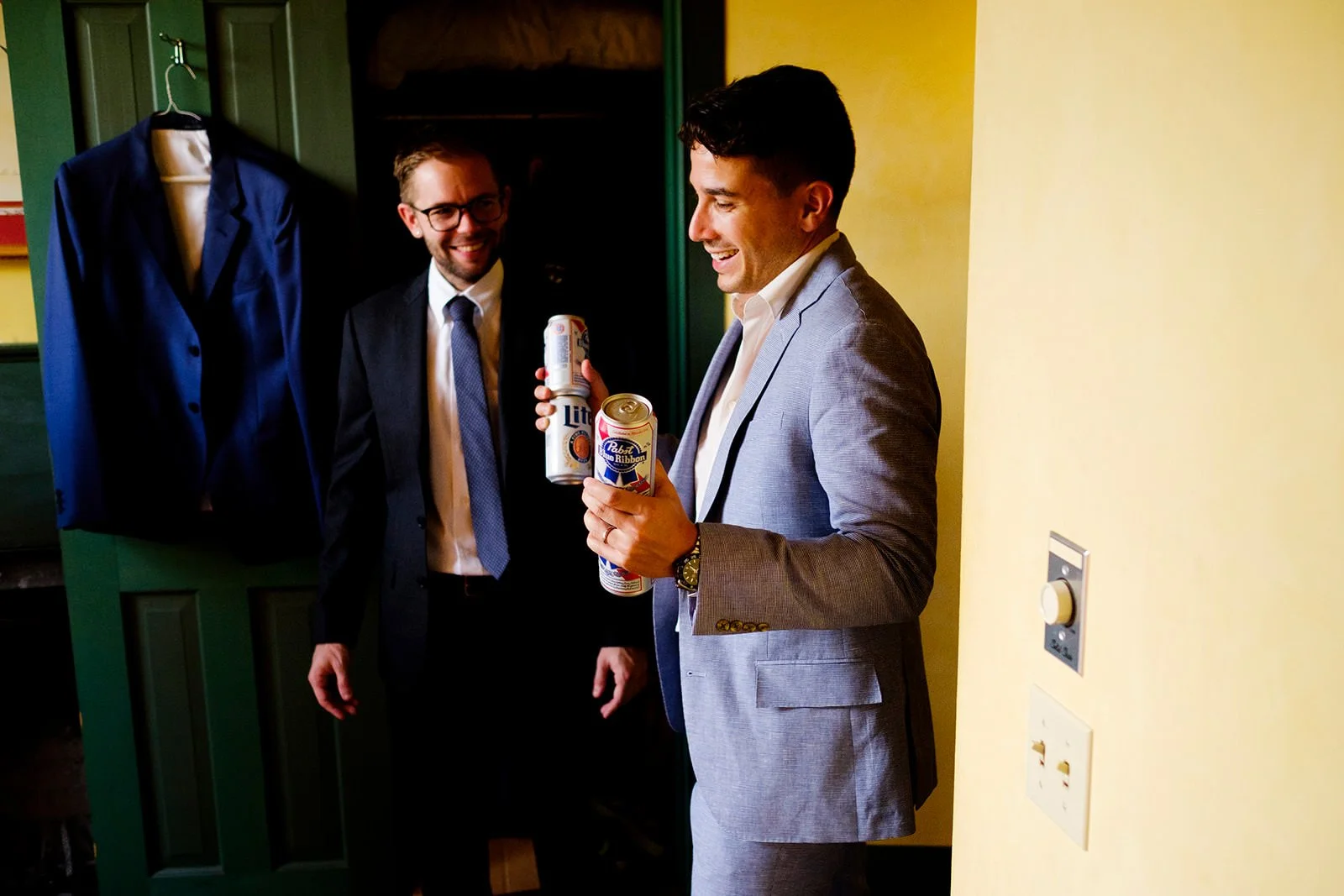 Two men in suits holding cans of beer, smiling and chatting in a room with a green door and a jacket hanging on a hook.