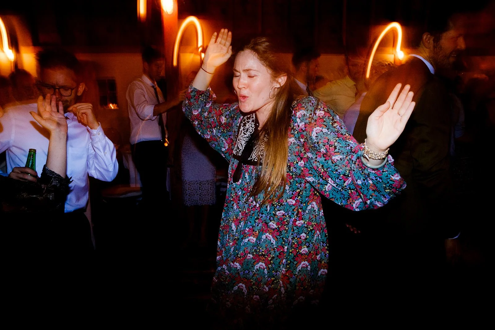 A woman dancing with her eyes closed and hands raised at a party, surrounded by other people in a dimly lit venue.