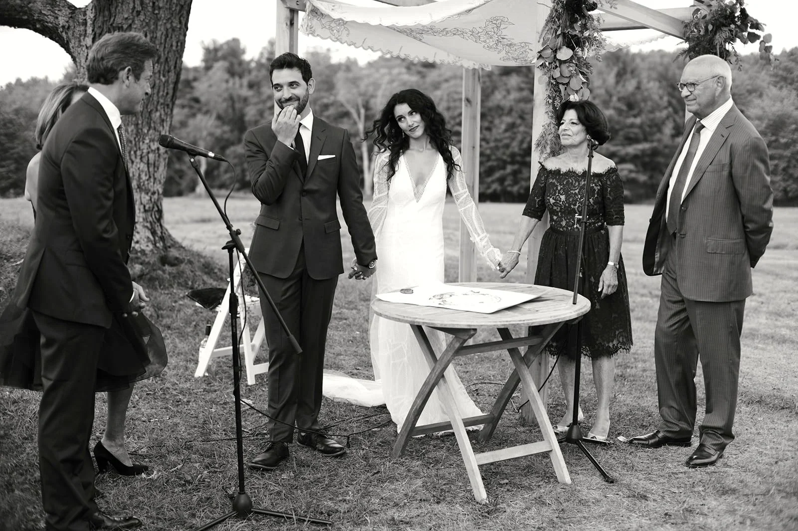 Black and white photo of a wedding ceremony outdoors with the bride and groom holding hands, surrounded by family and friends.