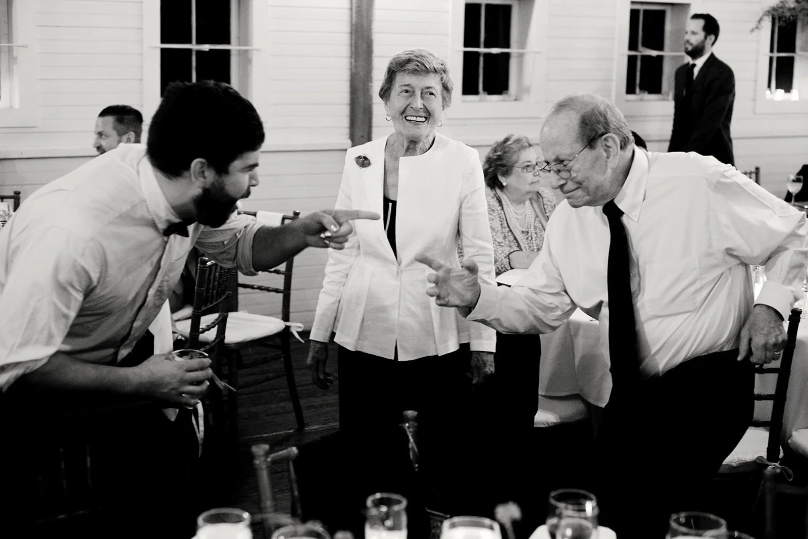 Black and white photo of four people at a social gathering, two men and two women. One man with a beard is leaning forward and pointing at another man who is smiling and leaning back slightly. The woman in the center is smiling and looking at the men