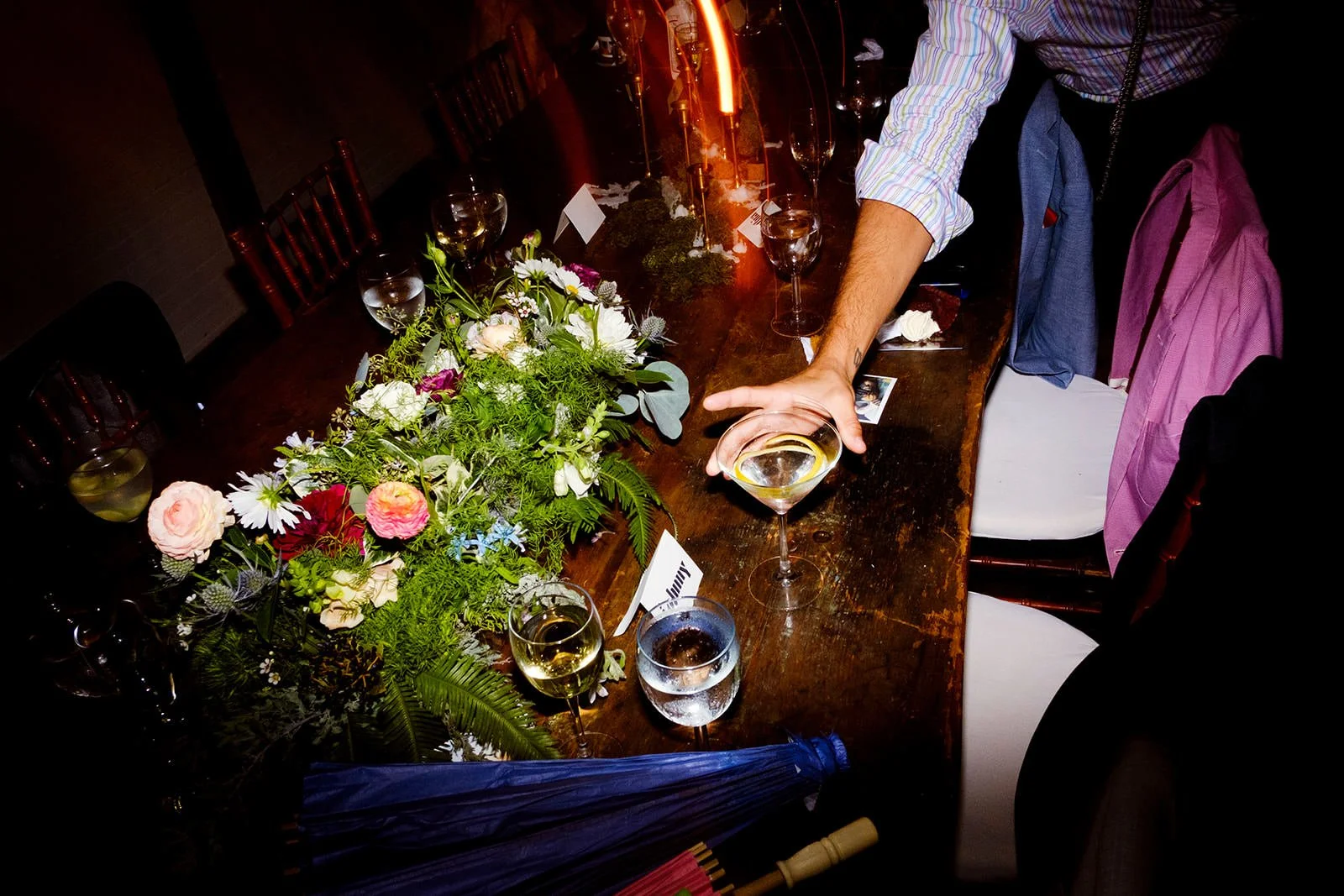 A long wooden table set for a celebration with a floral centerpiece, glasses of white wine, and a person's arm reaching for a glass. There are chairs around the table with some jackets draped over them, dim lighting, and a dark background.