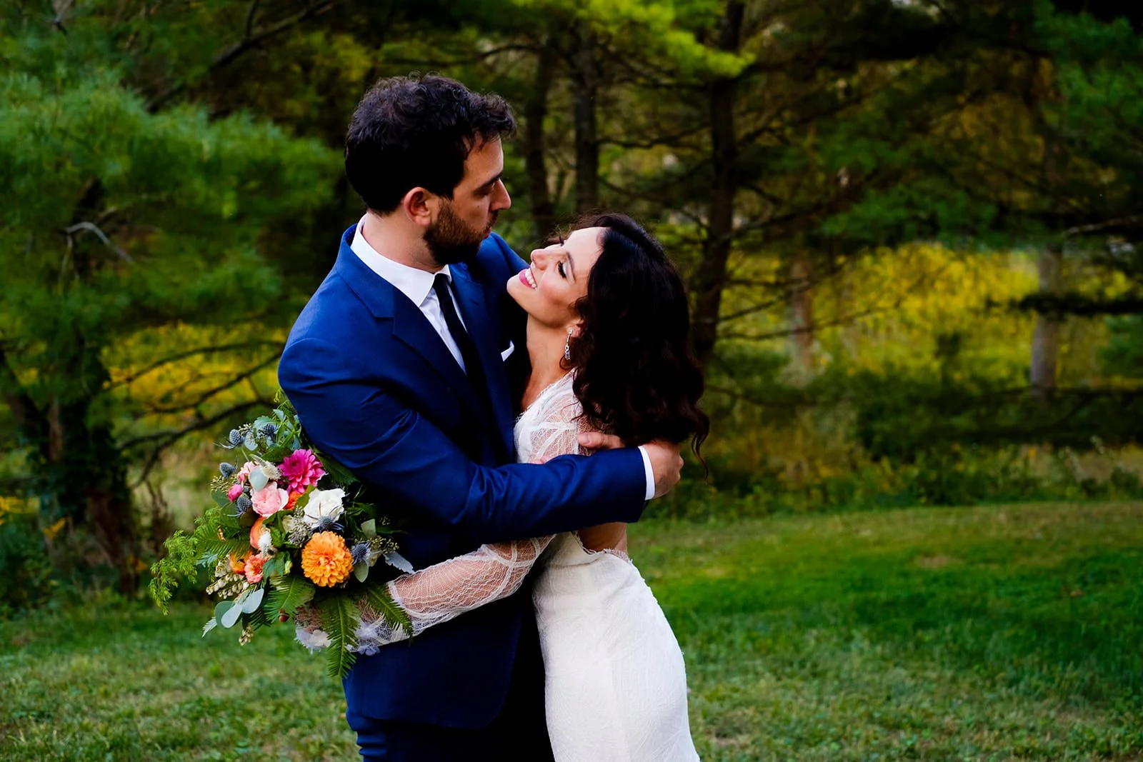 A bride and groom embrace outdoors in a grassy area with trees in the background. The groom is wearing a blue suit, and the bride is wearing a white wedding dress. The bride holds a colorful bouquet of flowers.
