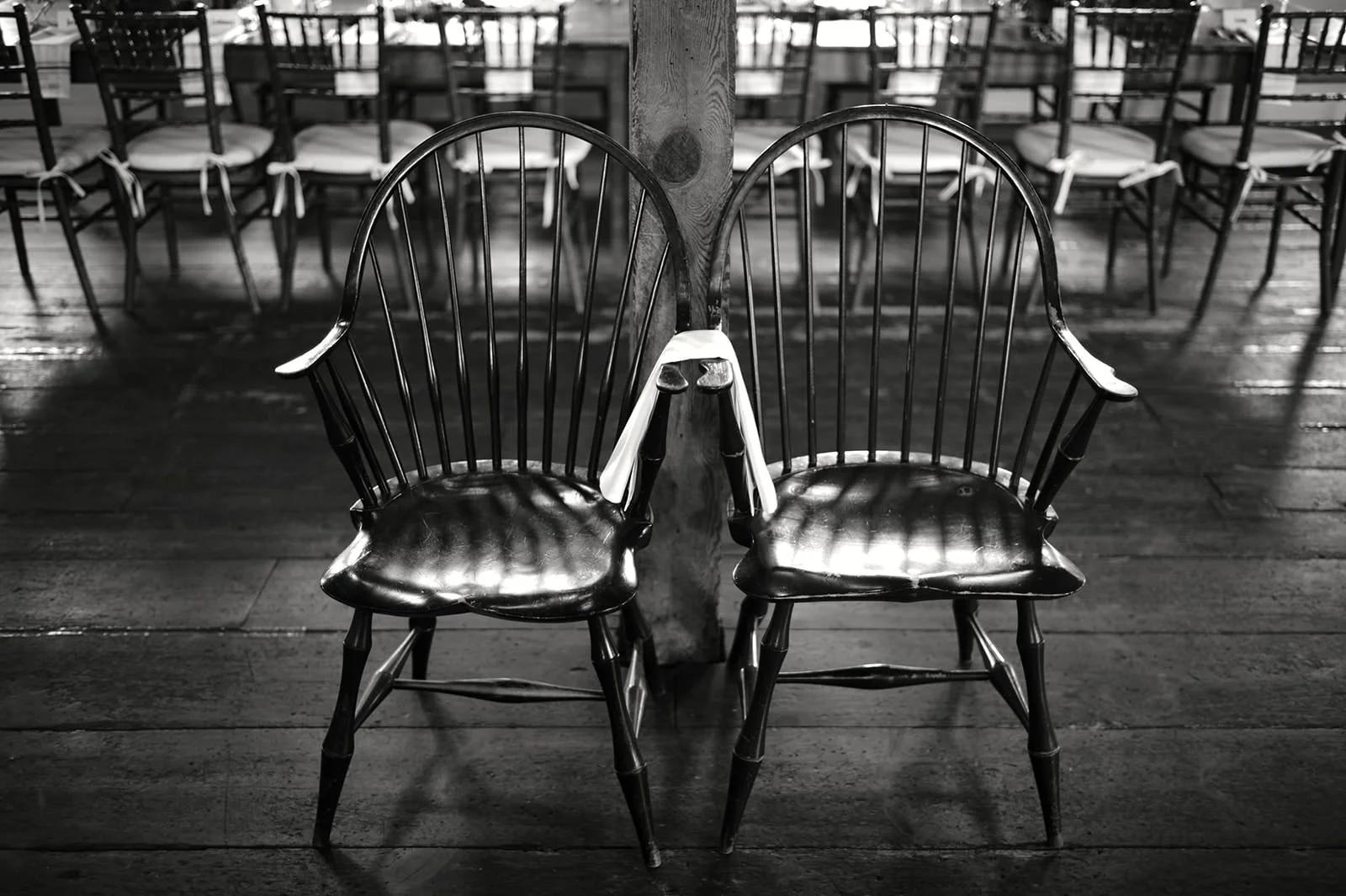 Two black wooden chairs with cushions set against each other, connected at the back by a wooden post, in a room with multiple rows of similar chairs in the background.
