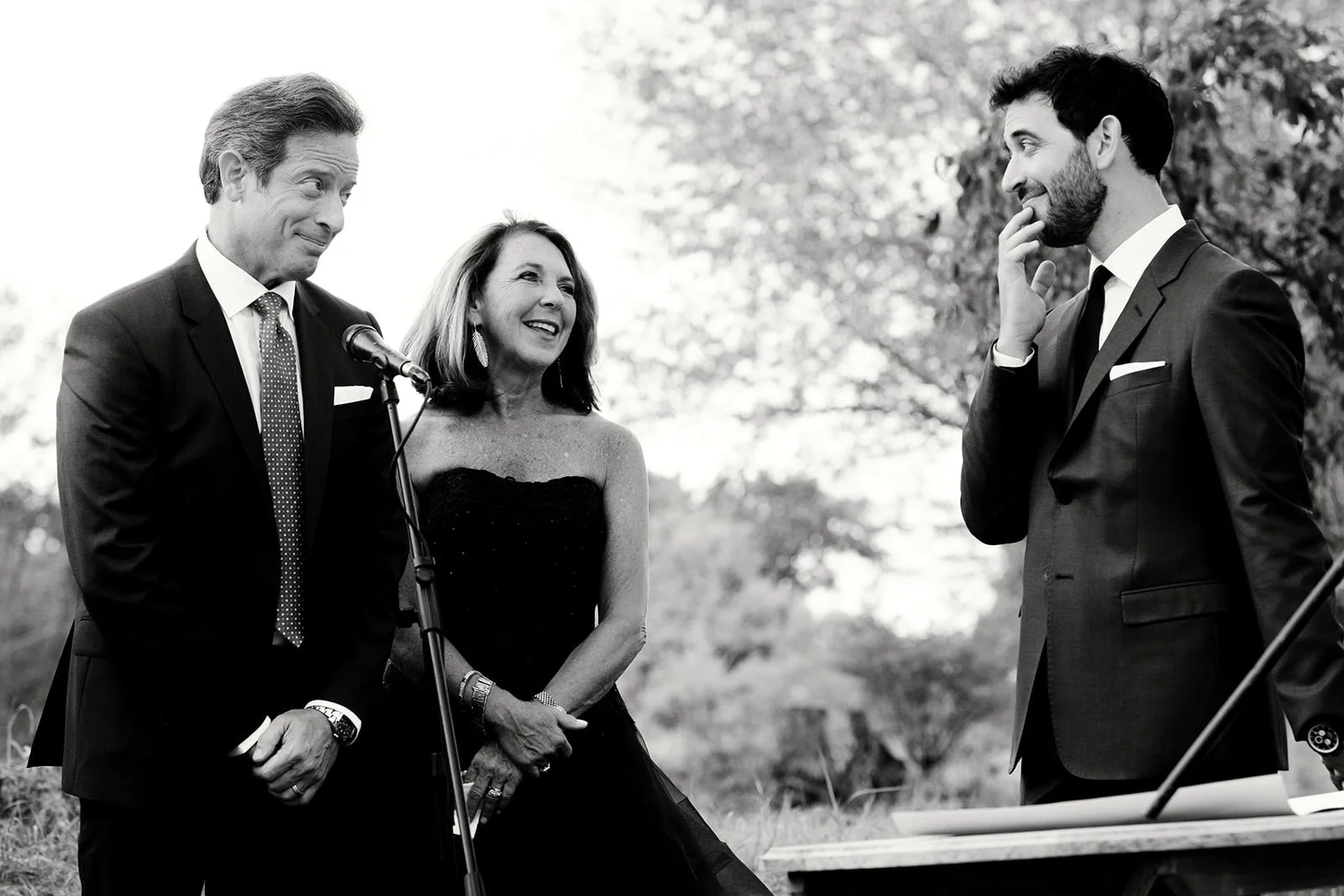 Black-and-white photo of three people outdoors at a formal event. A man in a suit and a woman in a strapless dress stand near a microphone, smiling and talking. Another man in a suit is on the right, smiling and touching his face while listening.
