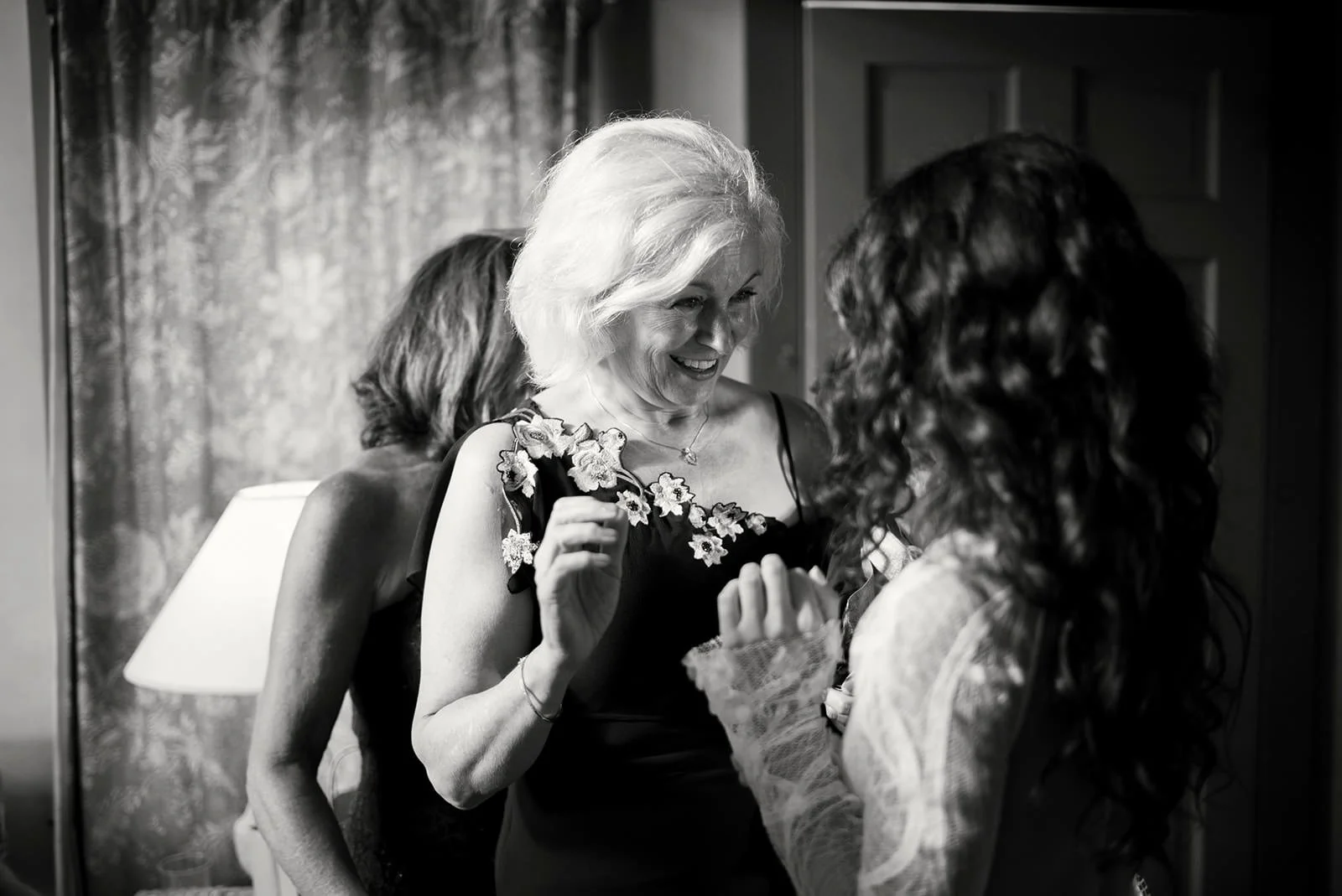 Two women, one elderly with light-colored hair and a floral dress, and one younger with dark curly hair in a lace top, share a joyful moment indoors. The elderly woman is smiling, and they are holding hands.