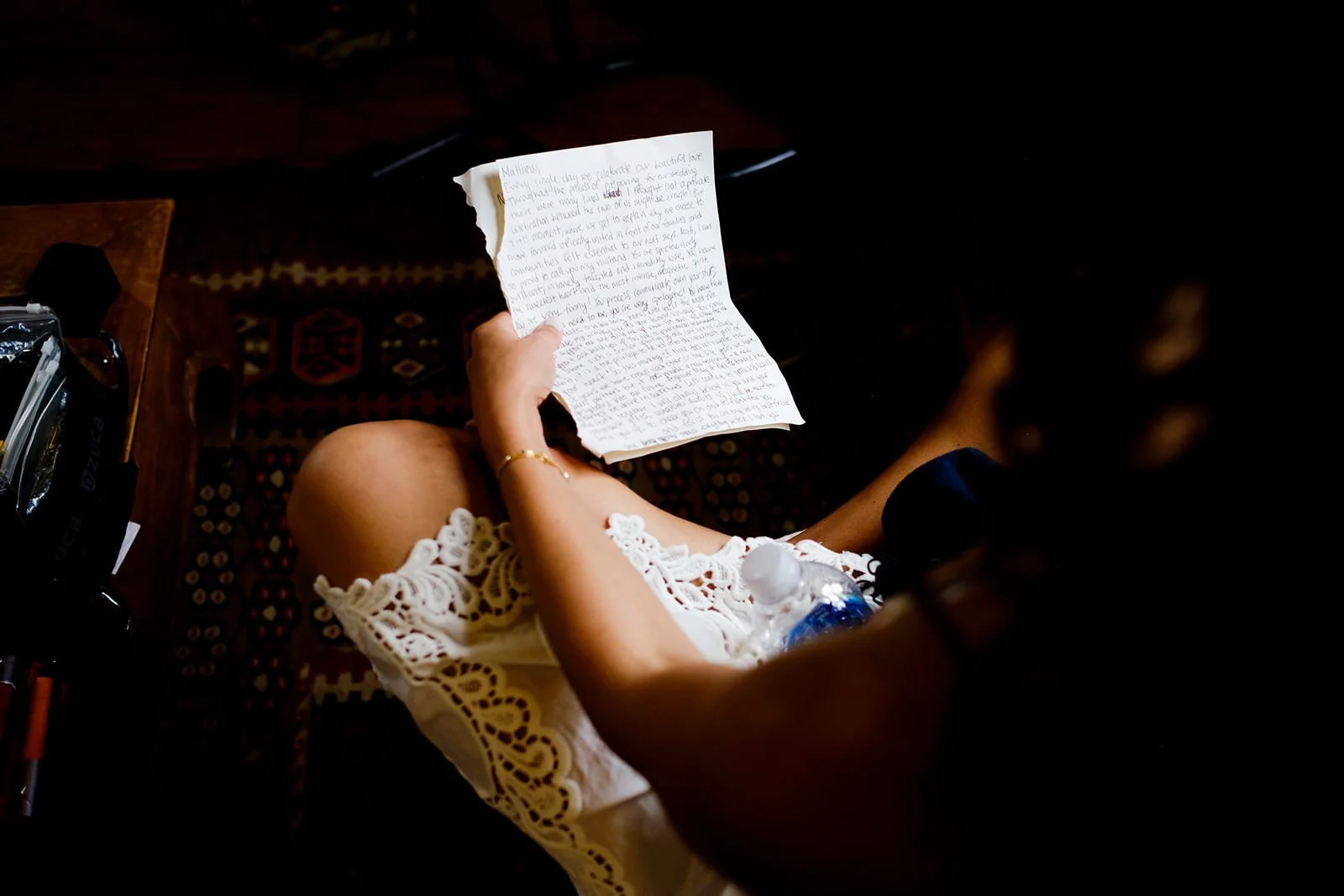 A woman is sitting on a patterned rug, wearing a cream-colored lace dress, holding a handwritten letter, with a water bottle on her lap and a plastic bag nearby.
