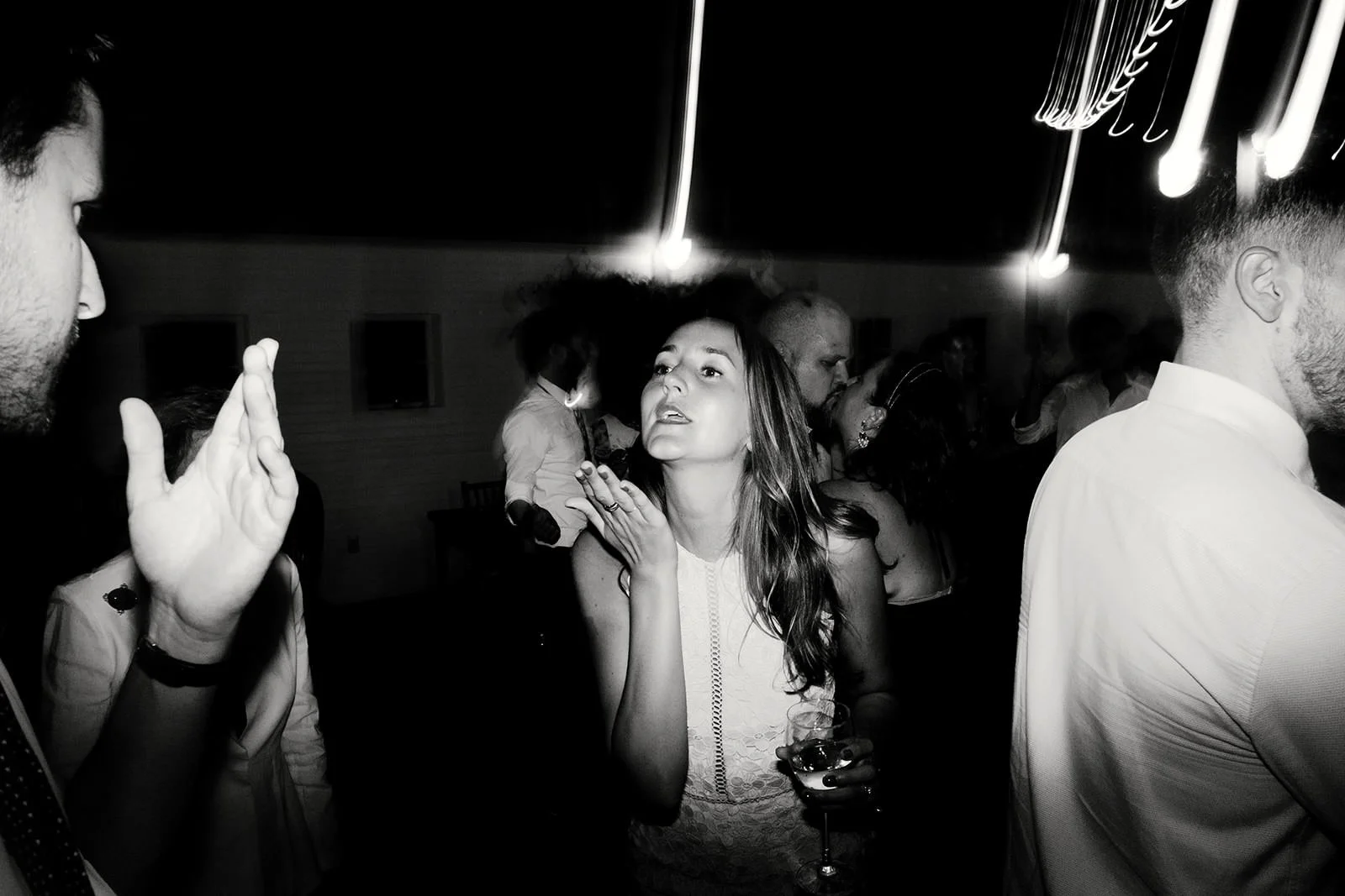 Black and white photo of a woman at a party, holding a glass of wine, with a slightly surprised or engaged expression, surrounded by other people dancing or socializing indoors.