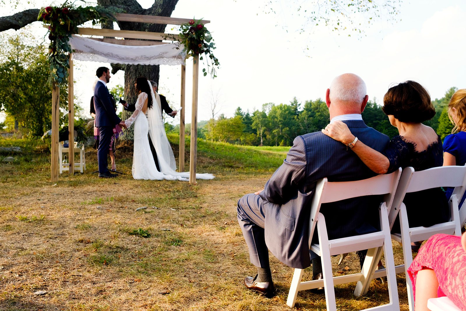 A couple getting married outdoors underneath a wooden arbor draped with white fabric and flowers, with family and friends seated on white folding chairs in a green, natural setting.