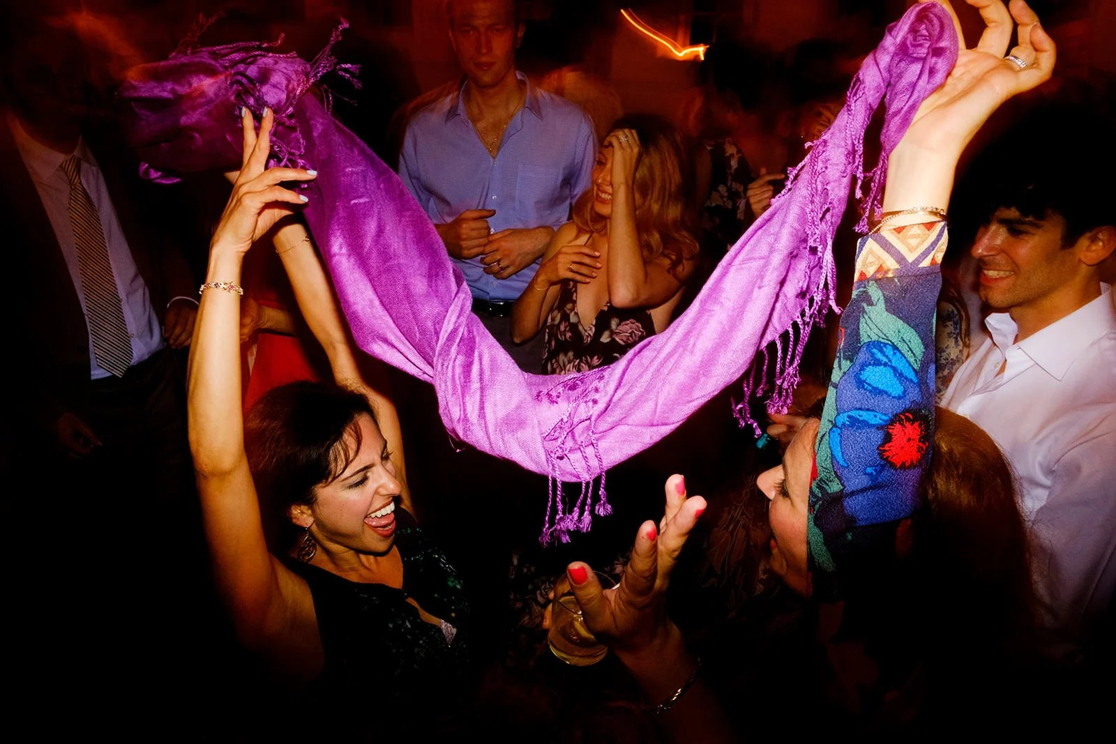 People celebrating at a party, lifting a woman dressed in purple in the air, with others smiling and enjoying themselves in the background.
