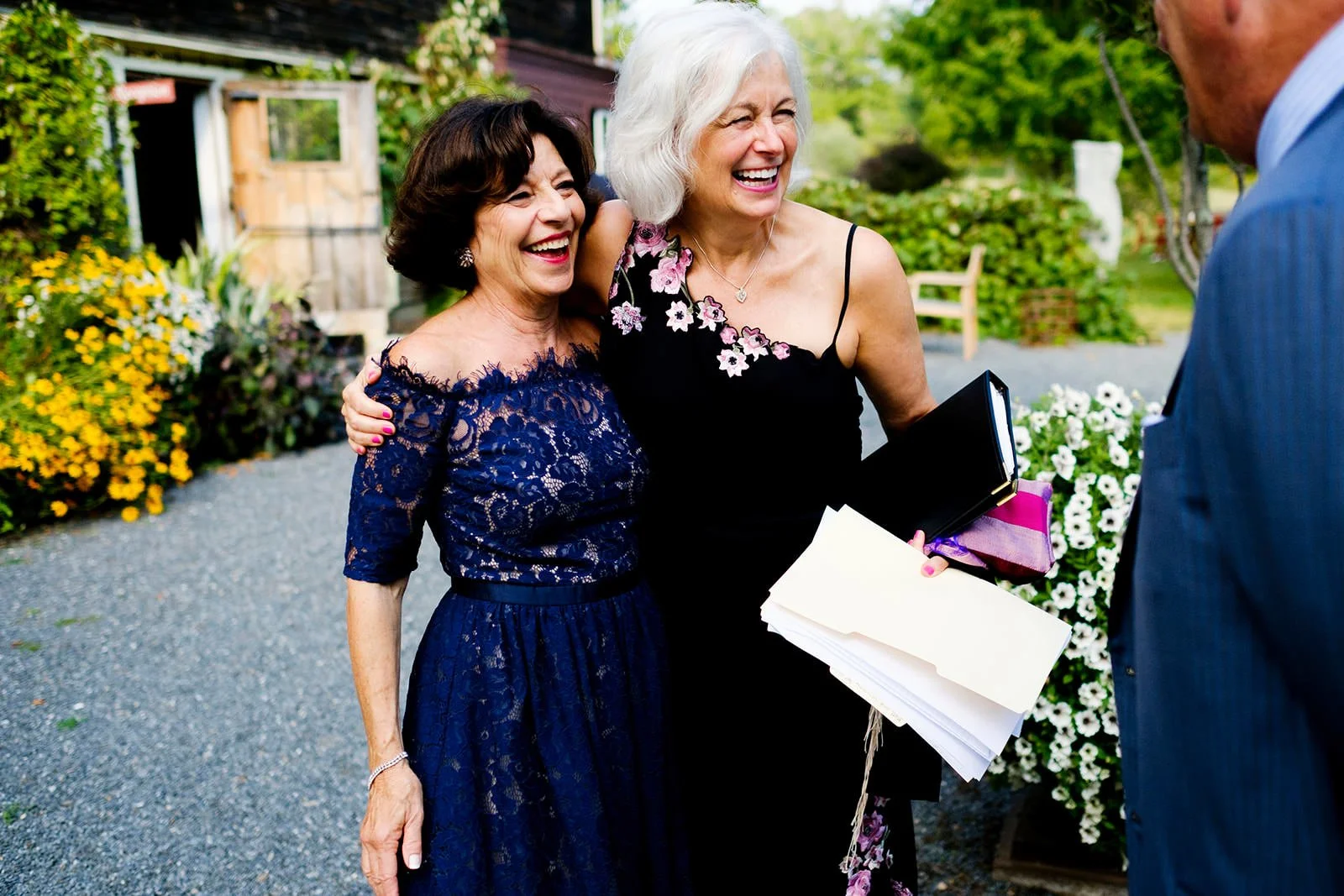 Two smiling women, one with dark hair in a navy lace dress and the other with white hair in a black dress with floral detail, standing outdoors with colorful flowers behind them, engaged in conversation with a man in a blue pinstripe suit.