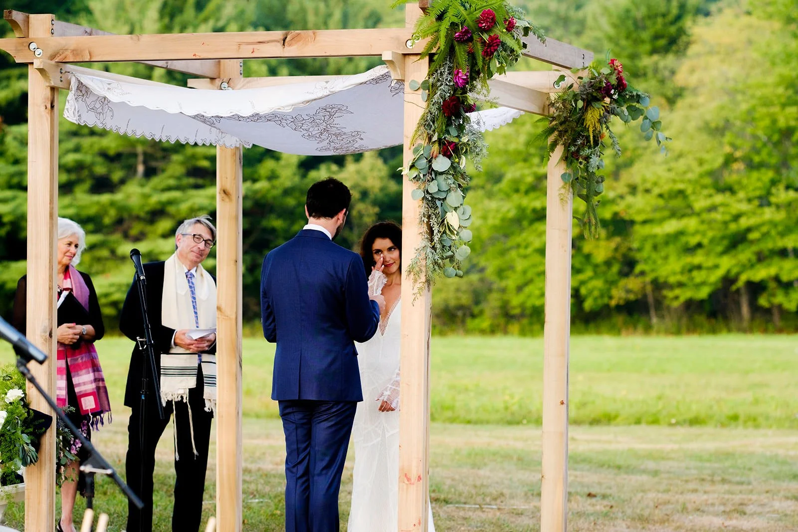 A couple getting married outdoors under a wooden arbor adorned with flowers and greenery, with two officiants and a woman beside them, set in a lush green park during daytime.