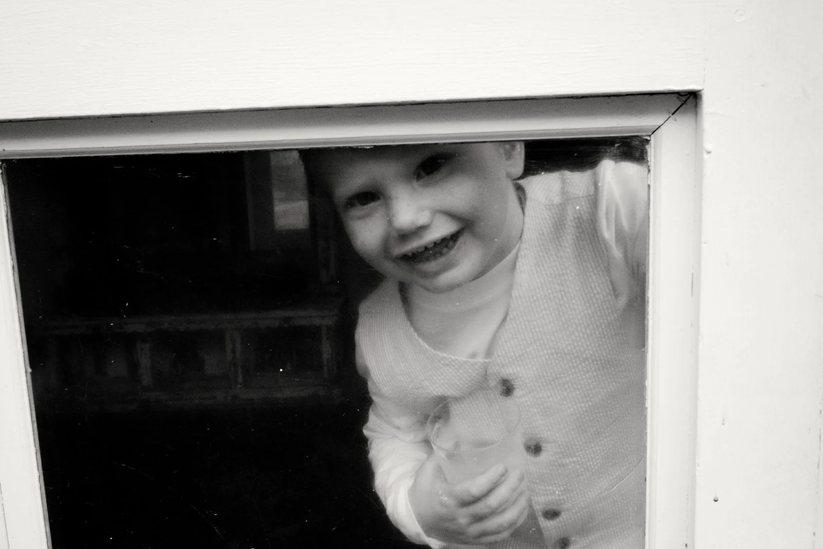 A young child smiling and looking through a small window in a door, holding a drink in one hand.