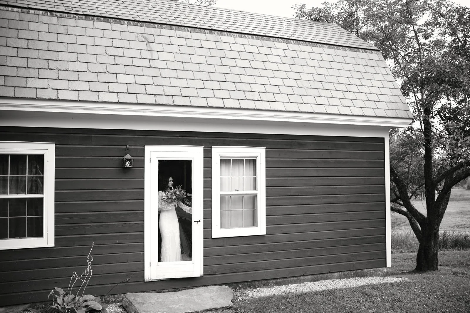 Black and white photo of a woman in a wedding dress standing behind a glass door of a house with horizontal siding and a gabled roof, holding a bouquet of flowers.