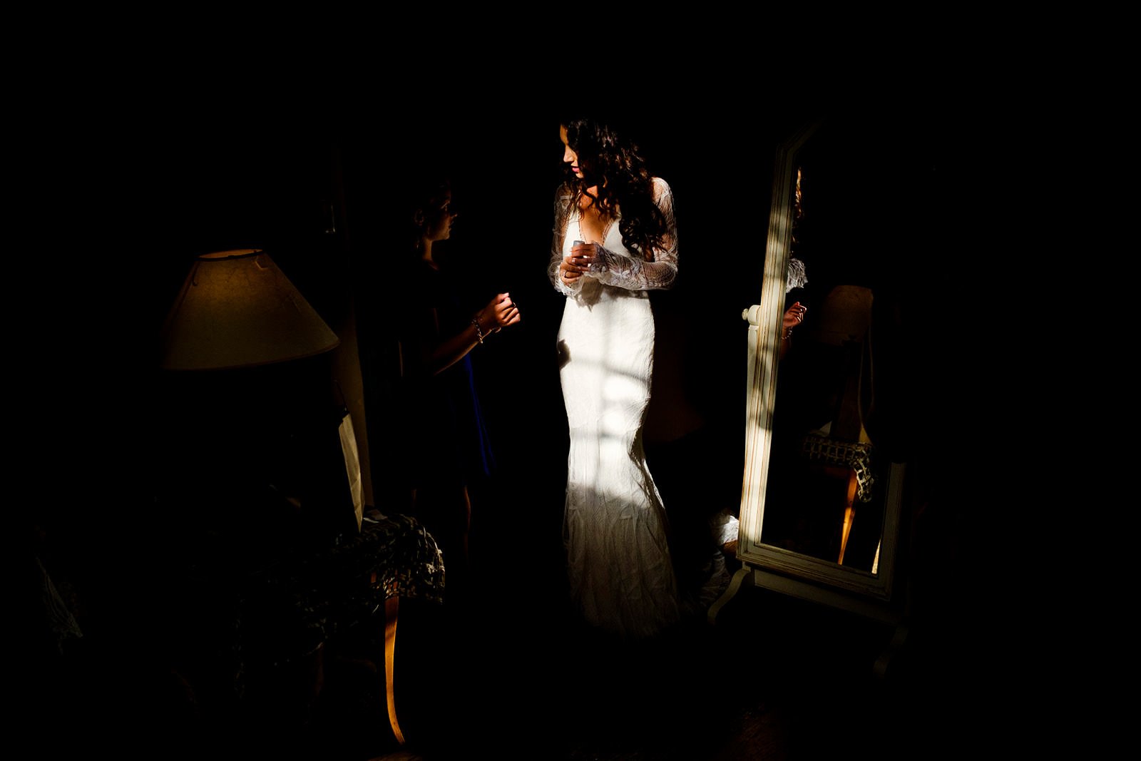 A woman in a white wedding dress standing in a dimly lit room, looking at a woman in a dark dress. The bride is holding a glass, and a mirror reflects her back. A lamp with a beige shade is on a table beside her.