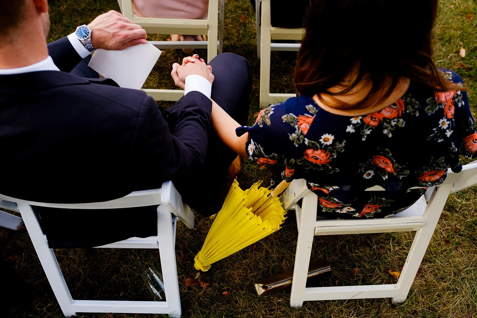A couple holding hands while sitting outdoors at a wedding, with a yellow umbrella resting on their chairs.