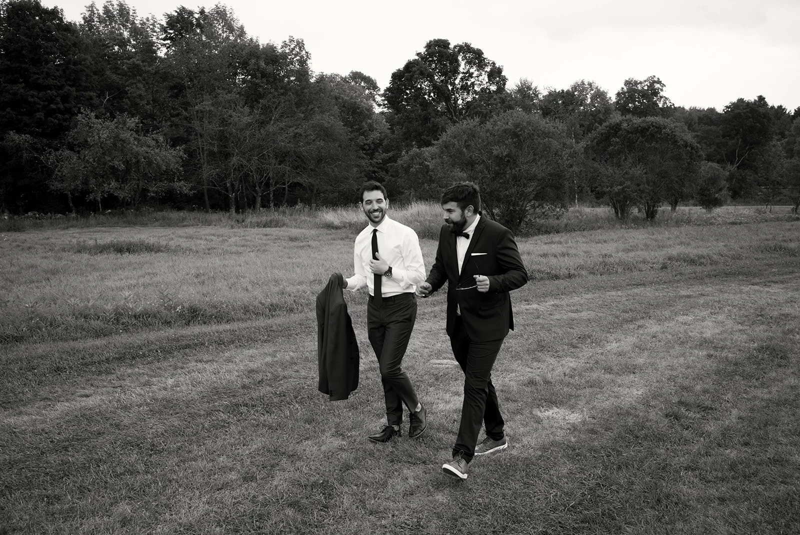 Two men in tuxedos walking across a grassy field with trees in the background, one holding a jacket and both smiling.